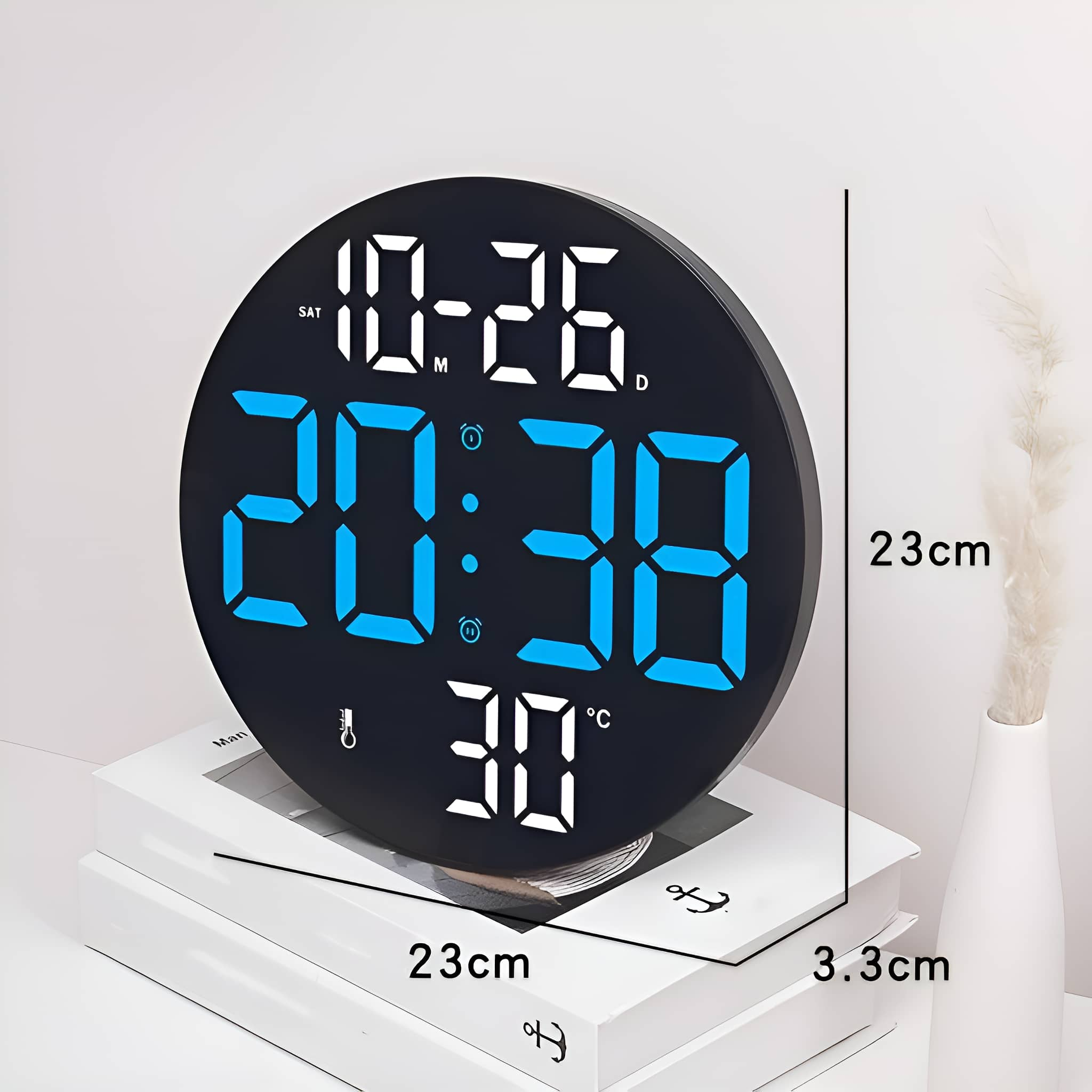 Eine schwarze Digitaluhr zeigt Zeit, Datum und Temperatur an. Sie steht auf weißen Büchern, rechts daneben eine Vase mit einer Pflanze.