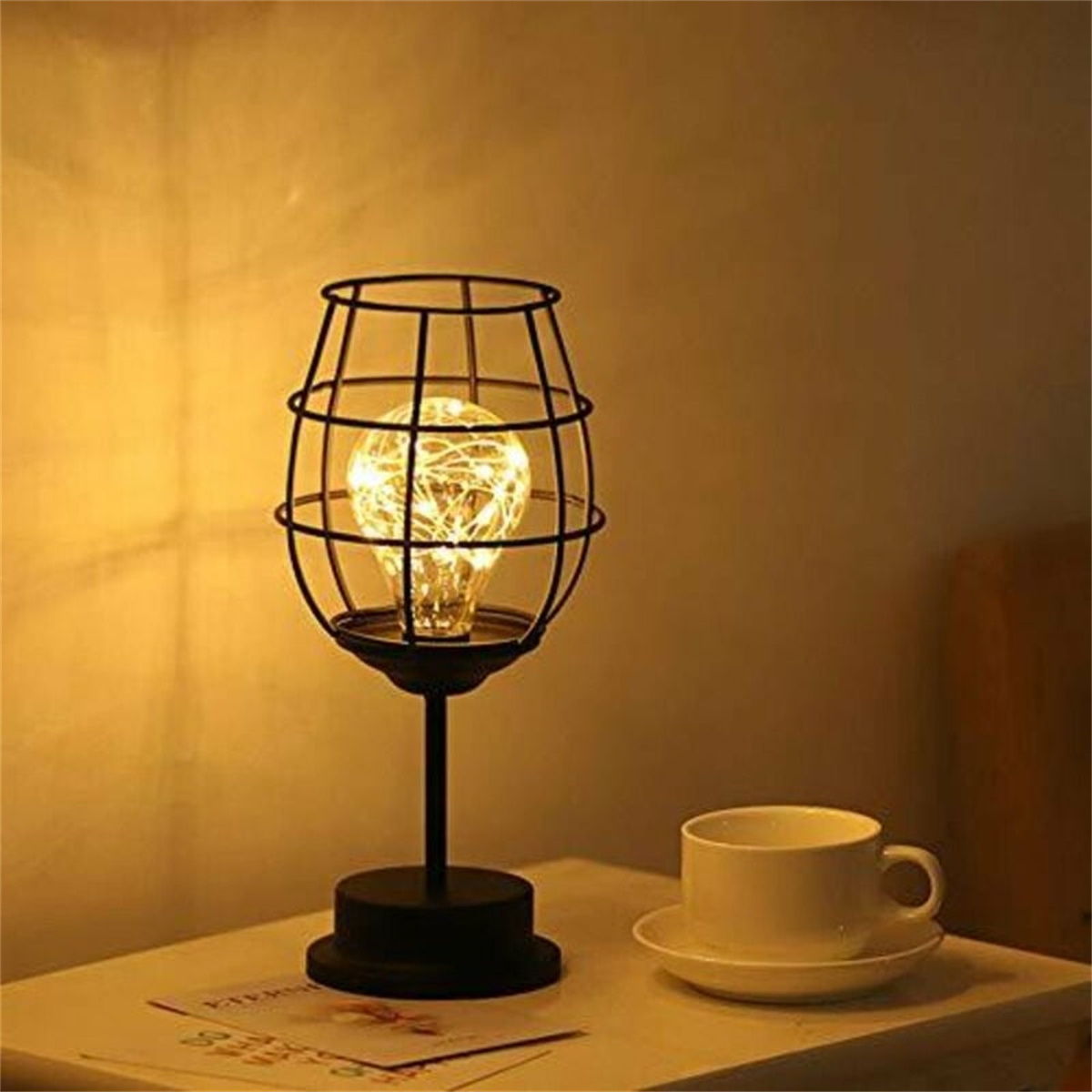 Eine schwarze Metalllampe mit käfigartigem Schirm, einer brennenden Glühbirne und einer weißen Tasse auf einer Untertasse. Warmes Licht.