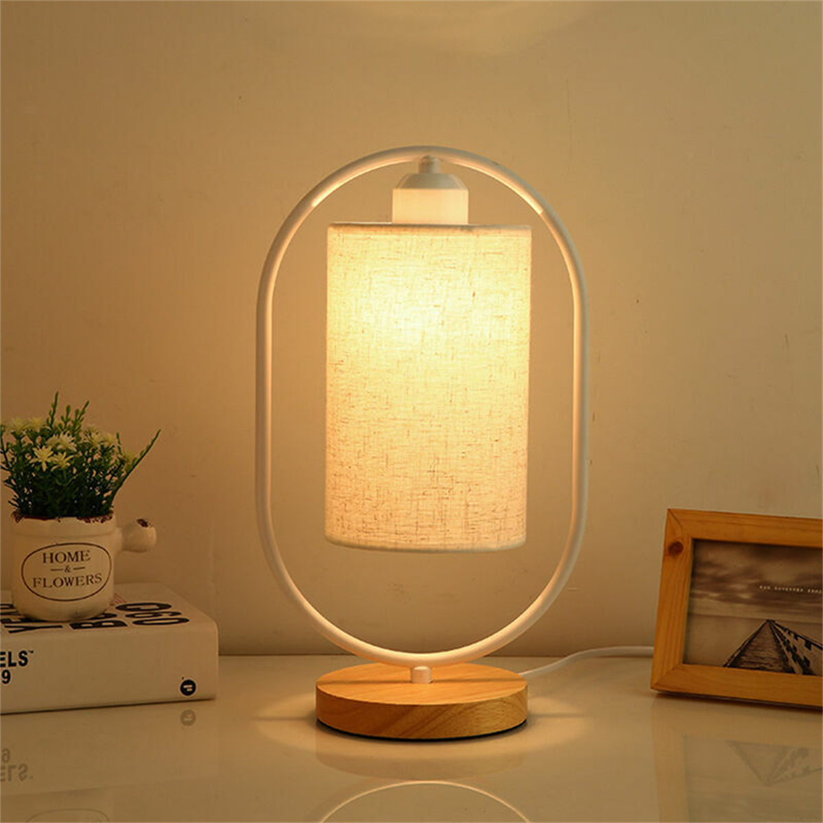 Eine moderne Tischlampe mit weißem Rahmen, beigem Schirm und Holzsockel. Eine kleine Pflanze und Bücher sind daneben.