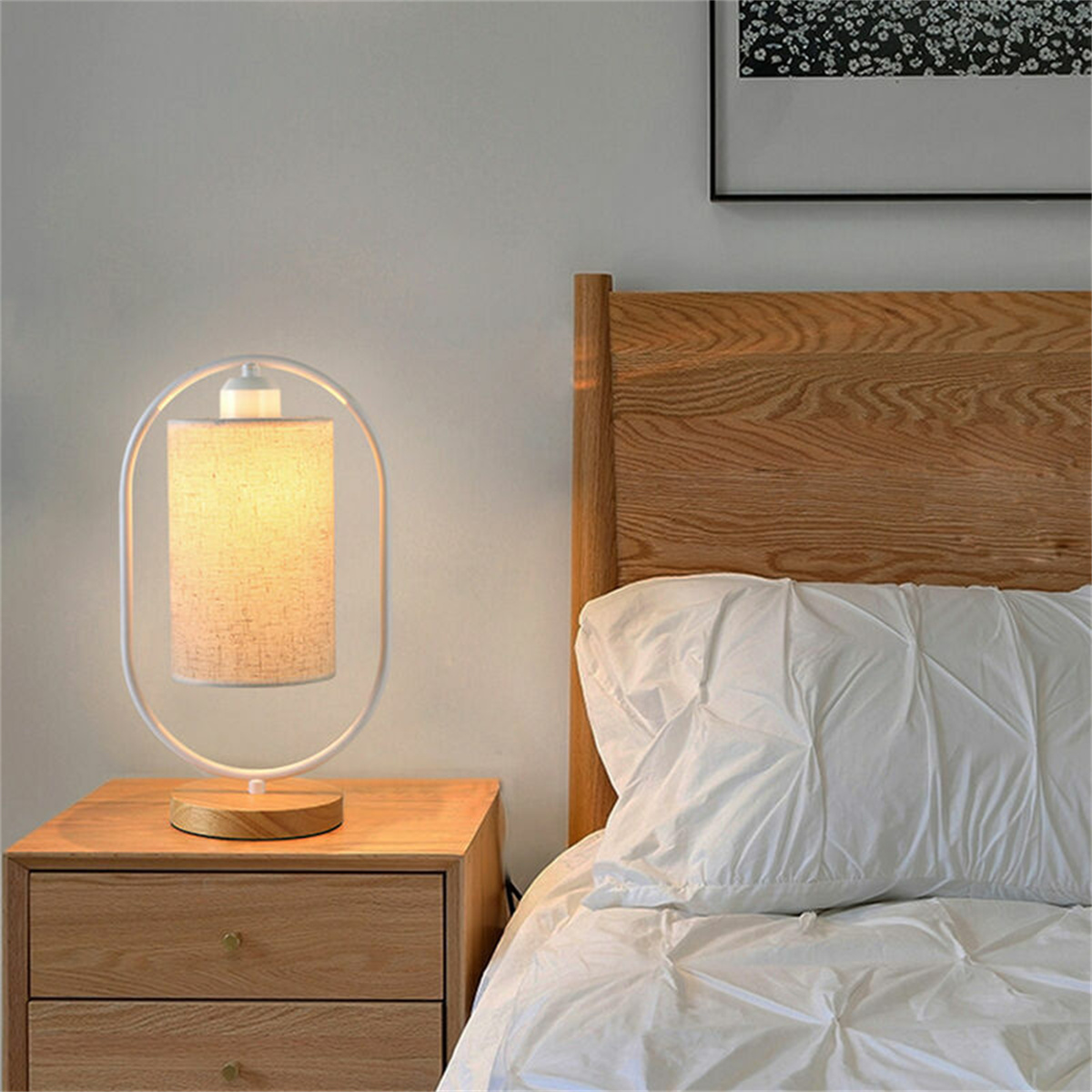 Tischlampe auf einem Nachttisch neben einem Bett. Die Lampe hat einen weißen Rahmen und einen beigen Schirm.