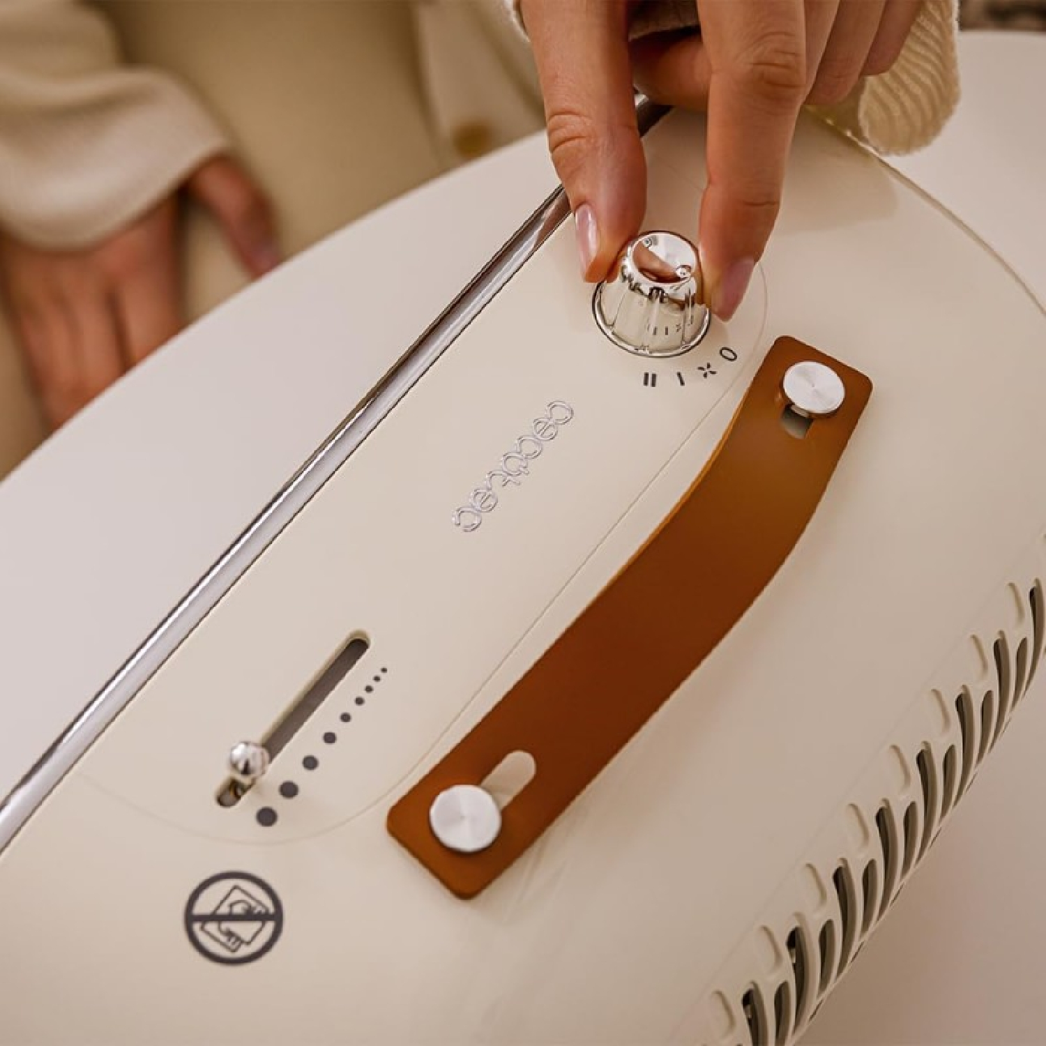 Una mano che regola una manopola su un ventilatore bianco e beige. Manico in pelle marrone e dettagli argentati visibili.