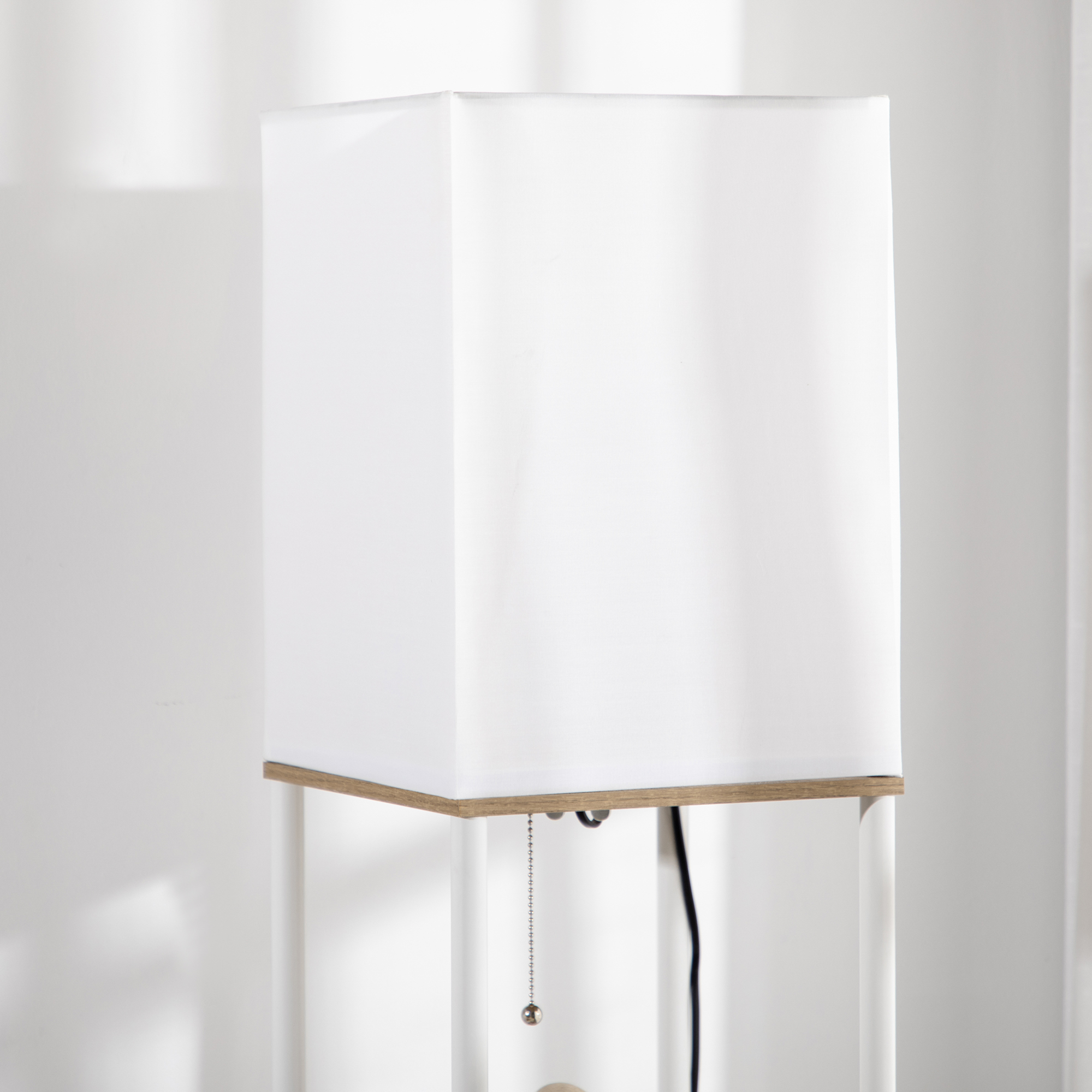 Una lámpara blanca con una pantalla cuadrada y una base de tono madera, sobre una pared blanca.