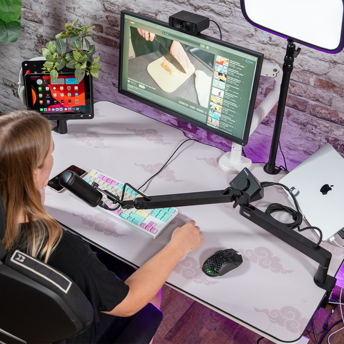 Frau an einem Schreibtisch mit Computer, Tablet, Mikrofon und anderem Streaming-Equipment. Hintergrund aus Ziegelmauer.