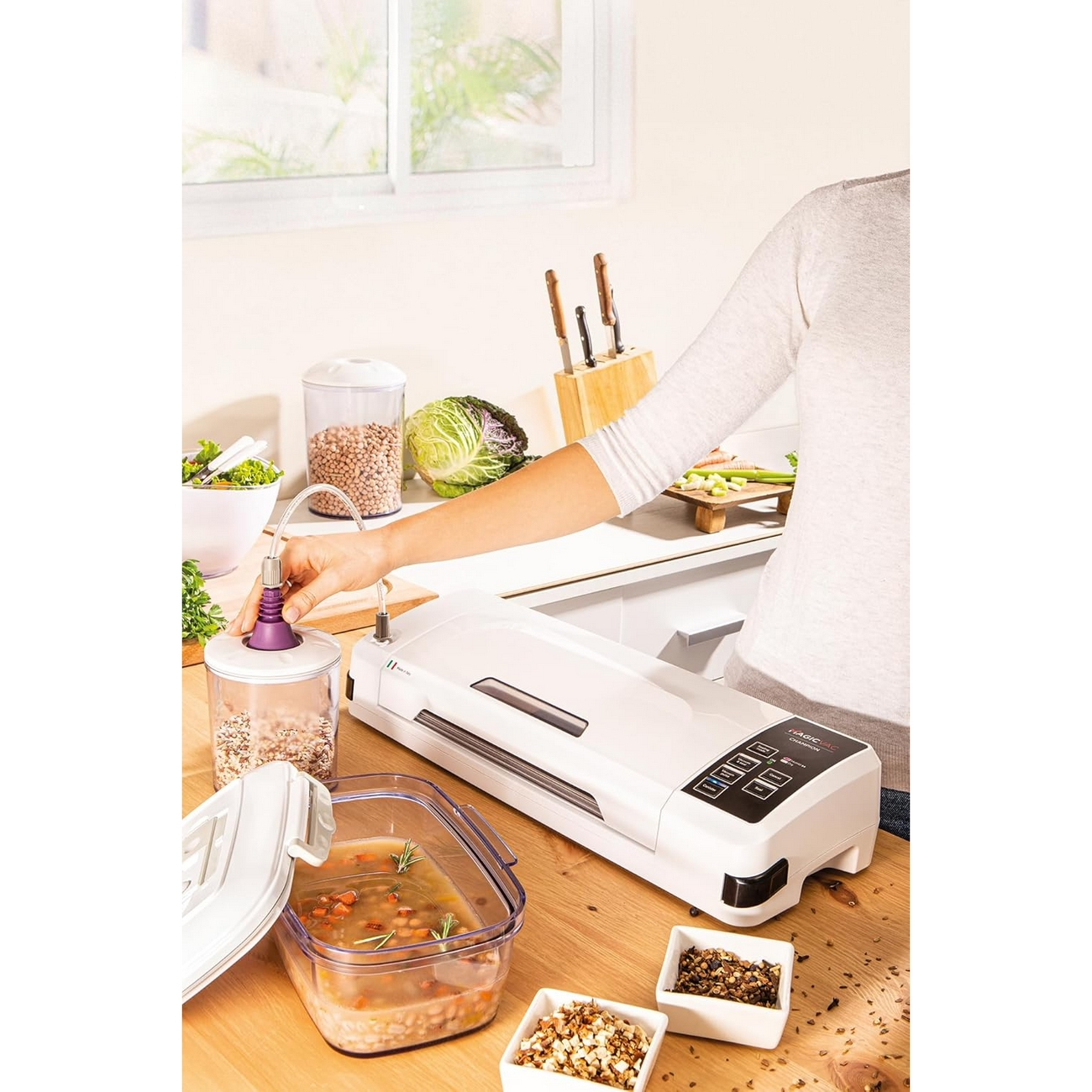 Scena kuchenna z kobietą używającą zgrzewarki próżniowej. Na stole jedzenie i pojemniki.