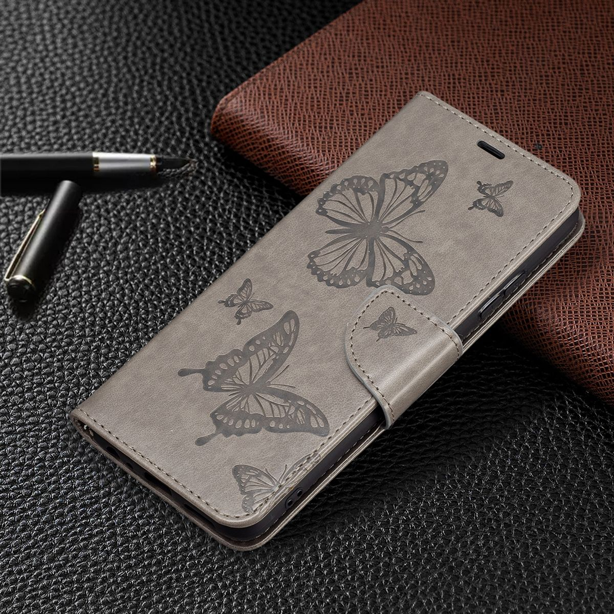 Graues Handycase mit Schmetterlingsdesign, auf dunkler Textur und braunem Portemonnaie.