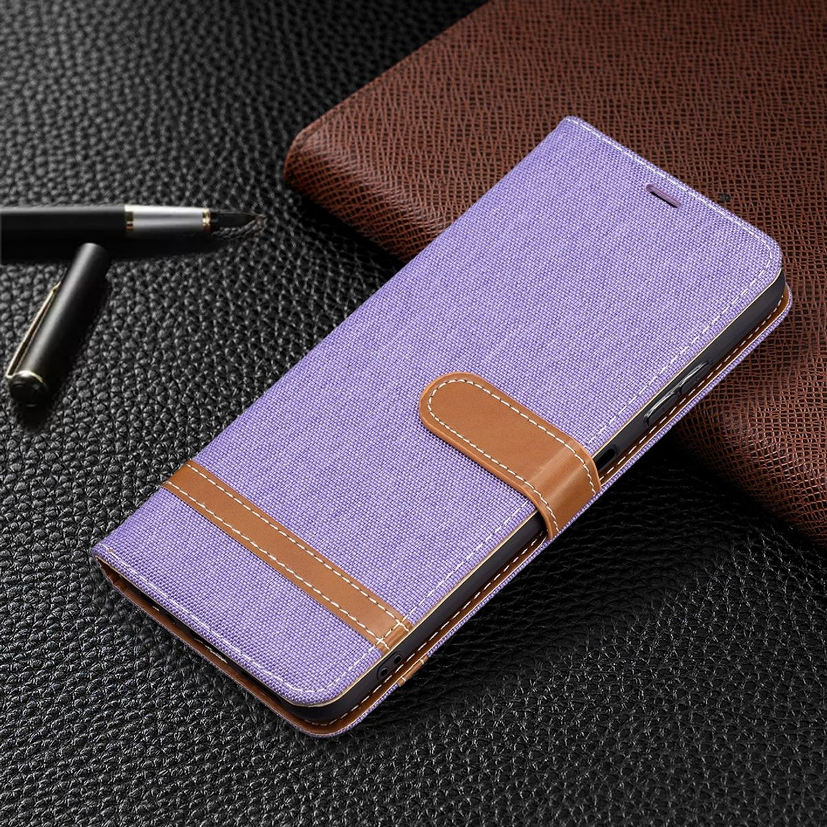 Eine lila Handyhülle mit braunem Riemen. Ein Stift liegt auf einer Lederoberfläche neben einer braunen Brieftasche.