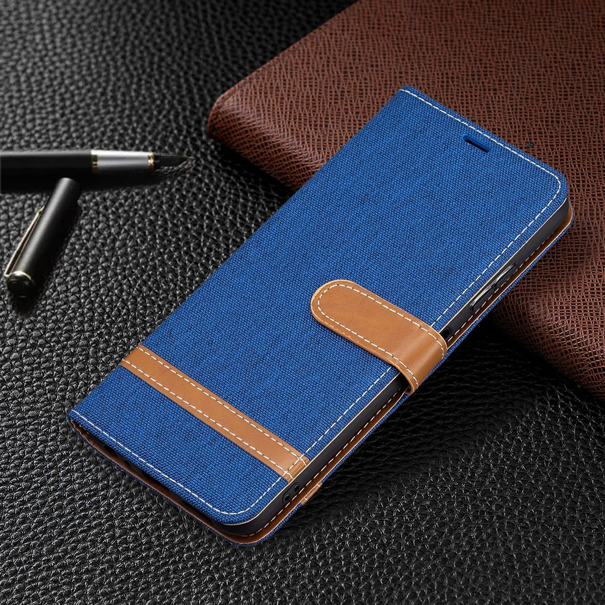 Blaue Handyhülle mit braunem Riemen und weißen Nähten auf strukturierter Oberfläche. Stift und Geldbörse im Hintergrund.