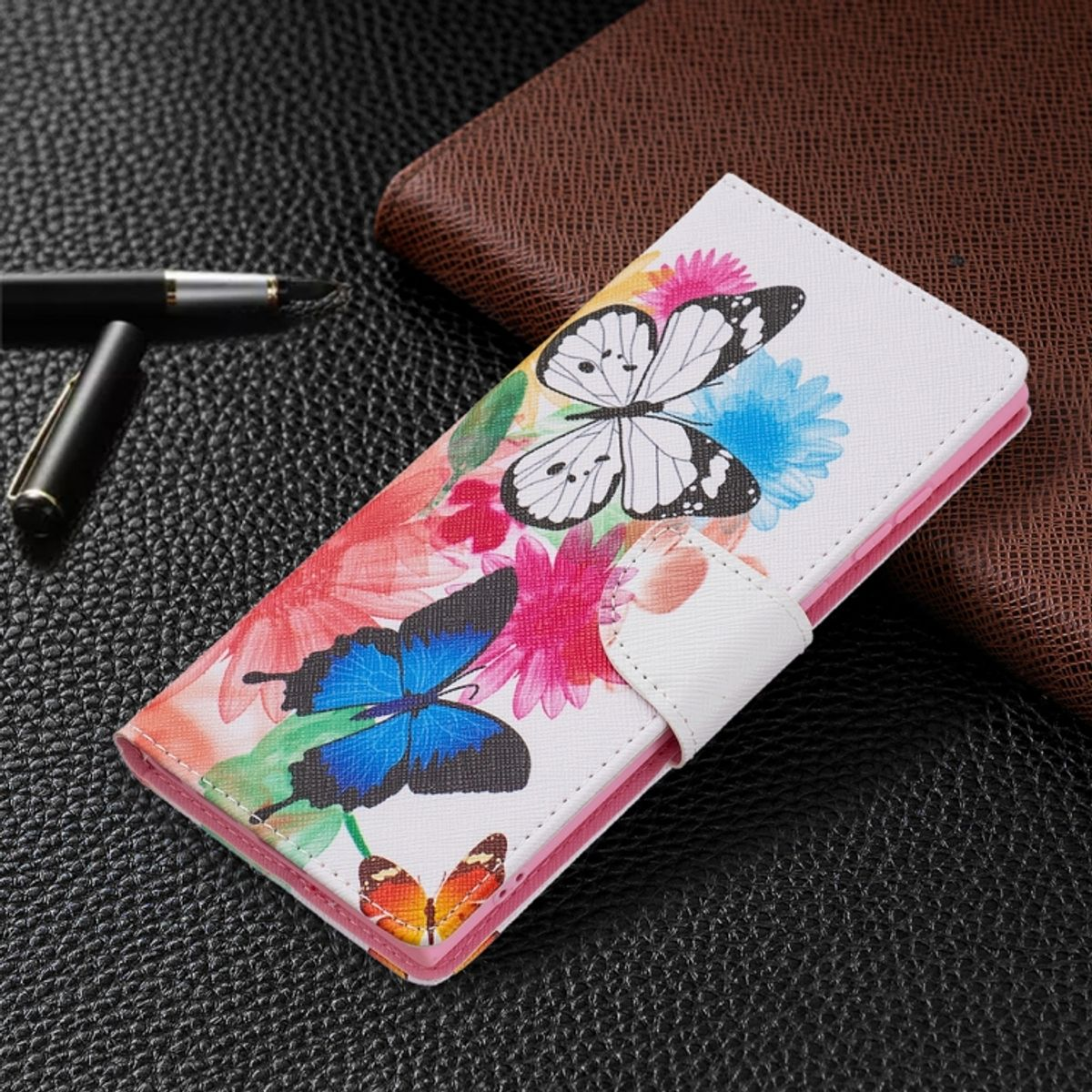 Weiße Handyhülle mit Schmetterlingen und Blumen. Stift und braune Geldbörse liegen auf dunkler Oberfläche.
