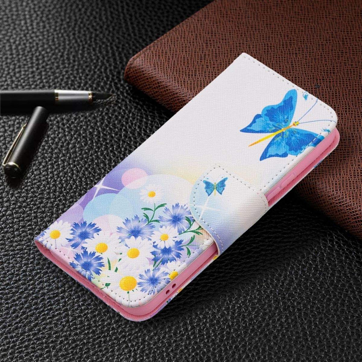 Weißes Handyhülle mit blauem Schmetterling und Blumen. Es liegt auf einer dunklen Oberfläche mit Stift und braunem Pad.