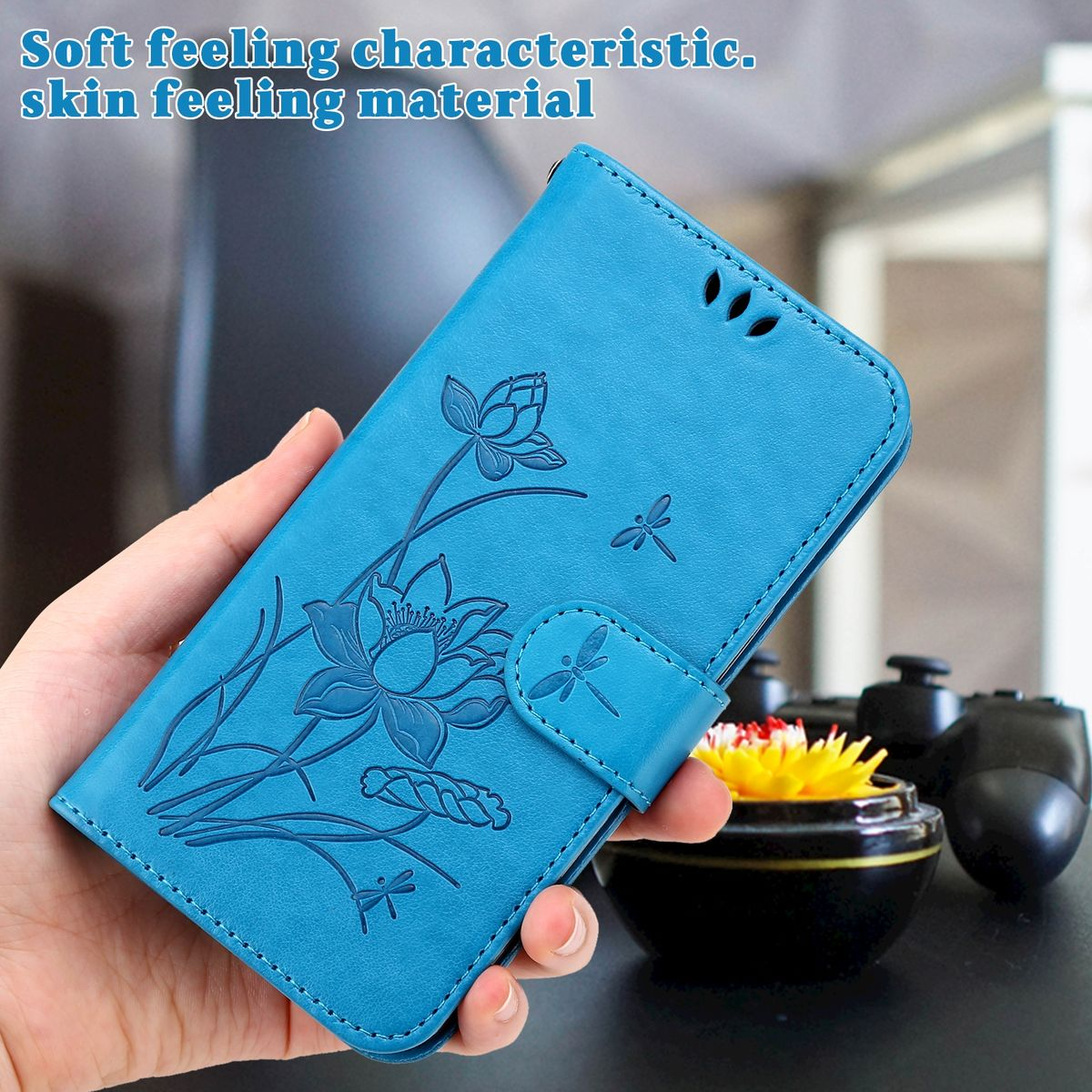 Blaue Handyhülle mit floralem Design, gehalten in einer Hand. Ein kleiner Topf mit einer Blume und ein Controller sind im Hintergrund.