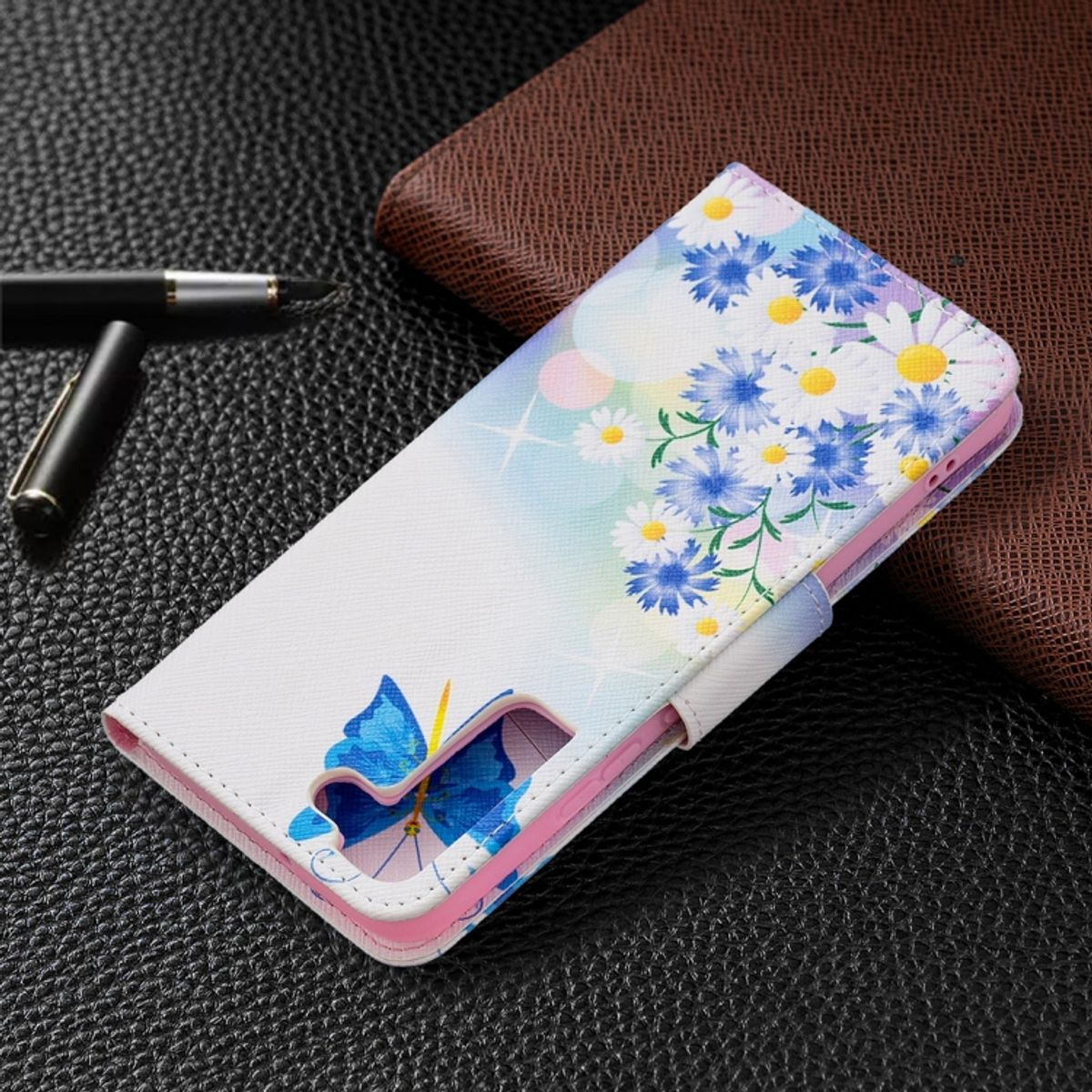 Handyhülle mit weißem Hintergrund, blauen Blumen und Schmetterlingen. Dunkle Lederoberfläche mit Stift.