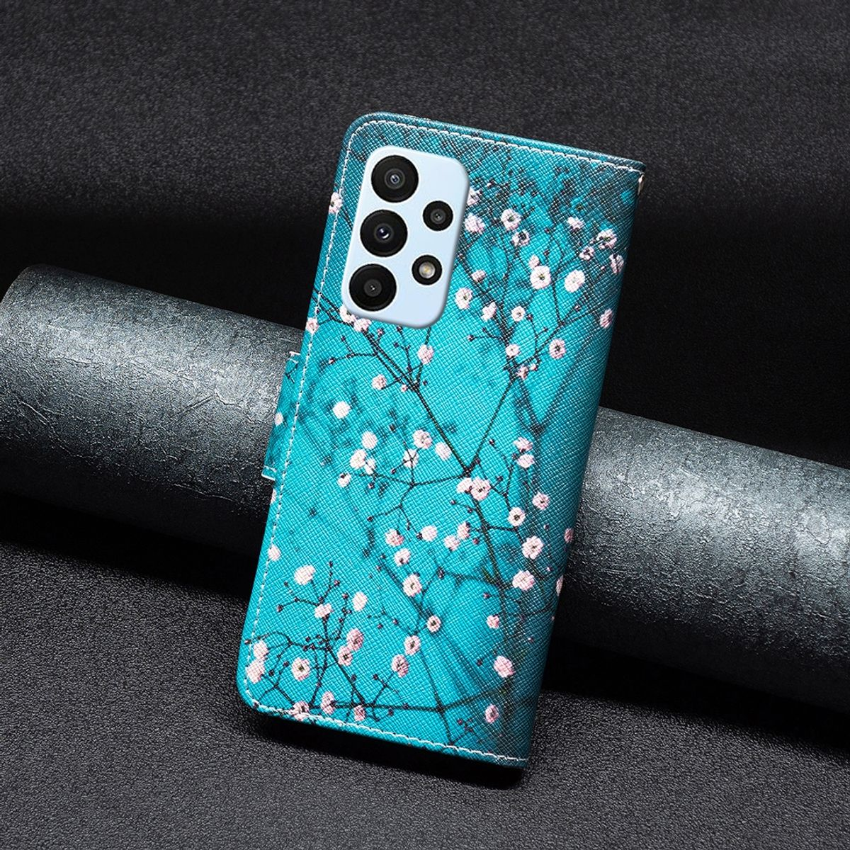 Blaue Handyhülle mit rosa Kirschblüten-Design. Handyhülle ist geschlossen. Dunkler Hintergrund mit einem Metallzylinder.