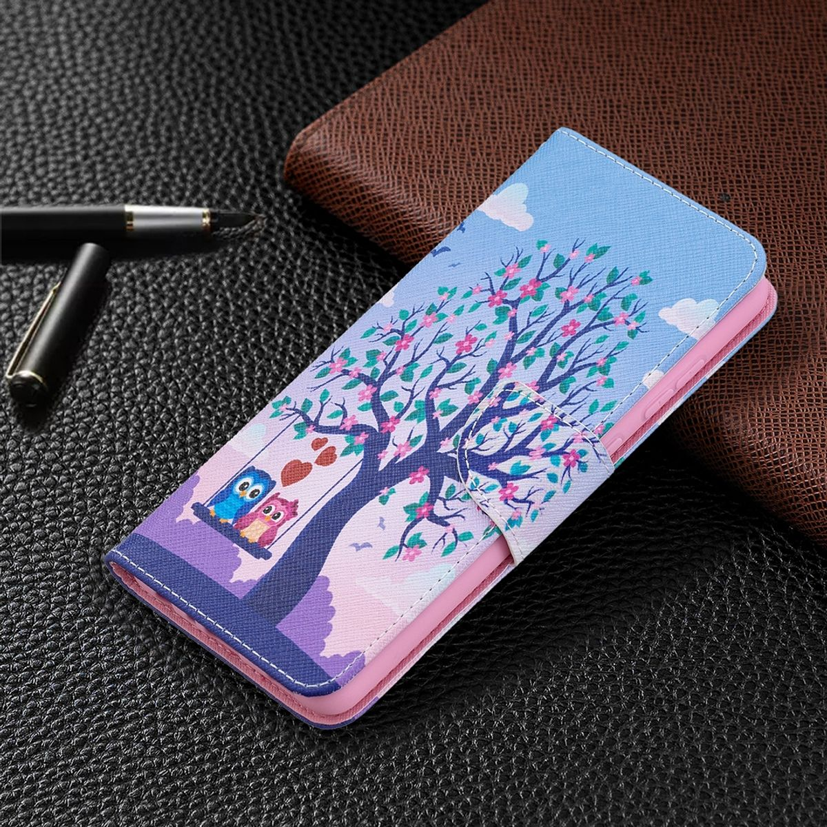 Blaue Handyhülle mit Baum- und Eulen-Design. Pinke und blaue Farben. Geöffnet, auf dunkler, lederartiger Oberfläche.