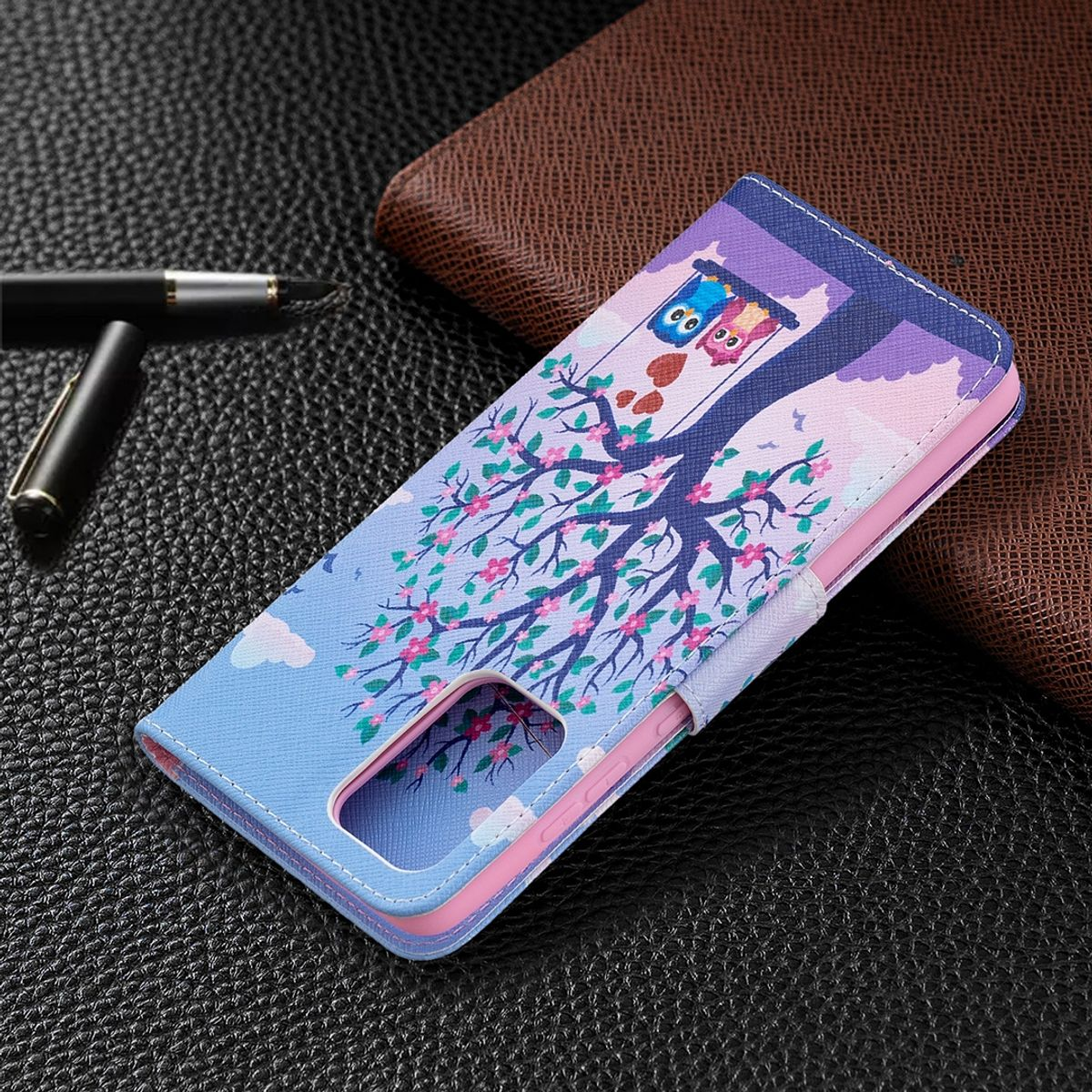 Blaue Handyhülle mit Eulen auf Schaukeln unter einem Baum. Brauner Lederhintergrund und Stift vorhanden.
