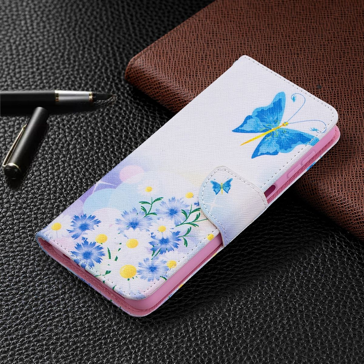 Weiße Handyhülle mit blauem Schmetterling- und Blumendesign. Auf einem Tisch neben einem Stift und einem braunen Portemonnaie.