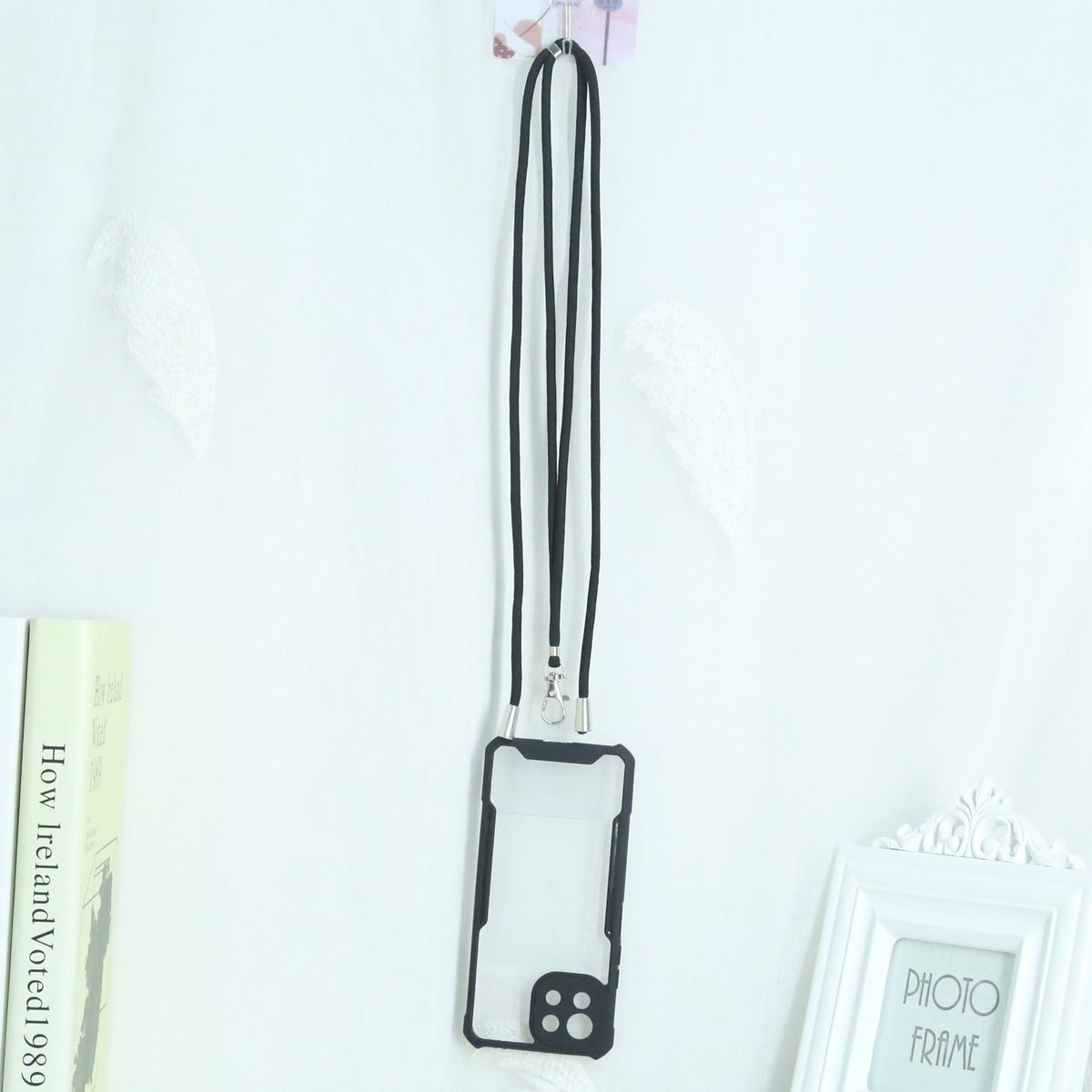 Schwarze Handyhülle mit schwarzem Band. Hängt an einer weißen Wand. Ein Buch und ein Rahmen links.