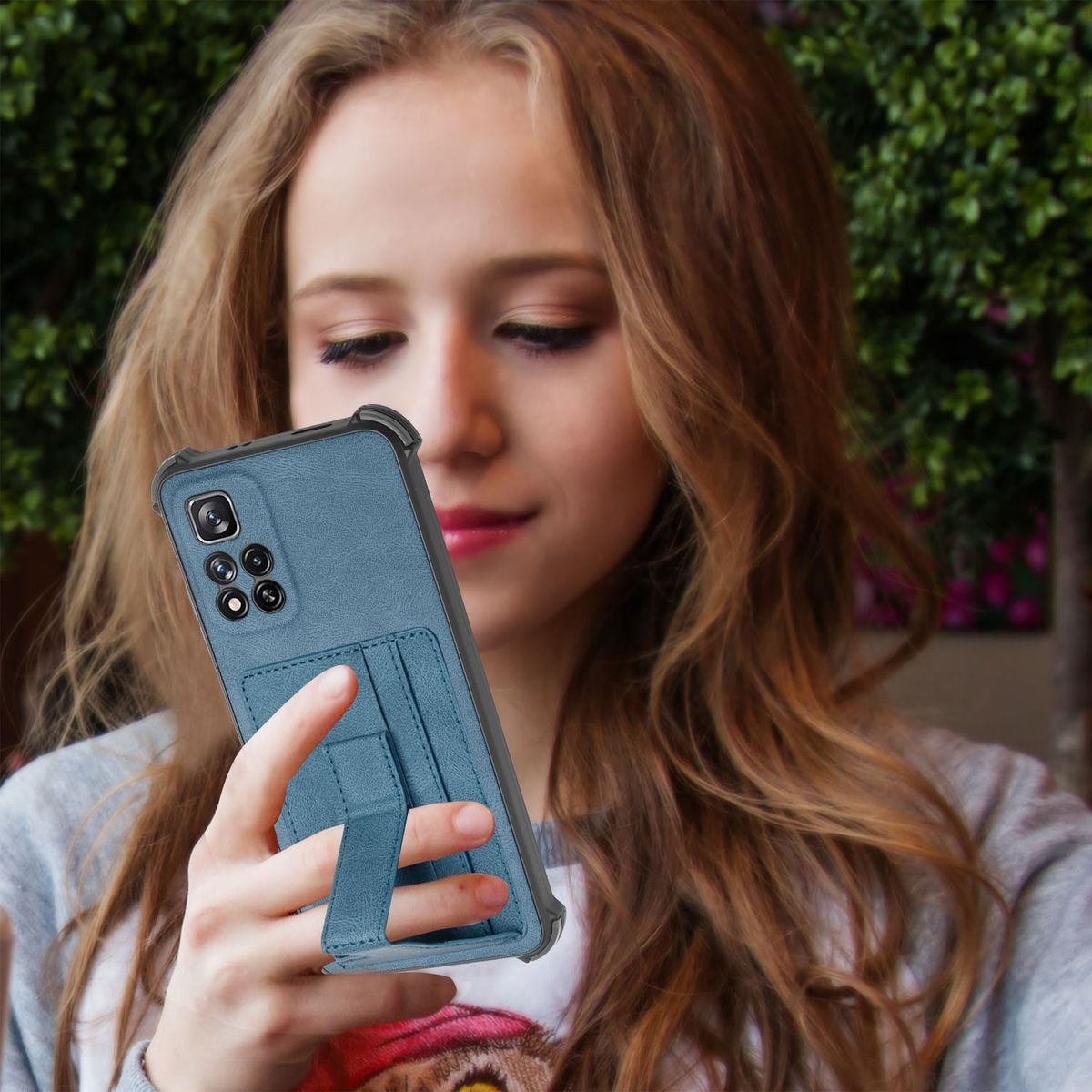Eine Frau hält ein Telefon in einer blauen Hülle. Der Hintergrund ist mit grünen Pflanzen verschwommen.