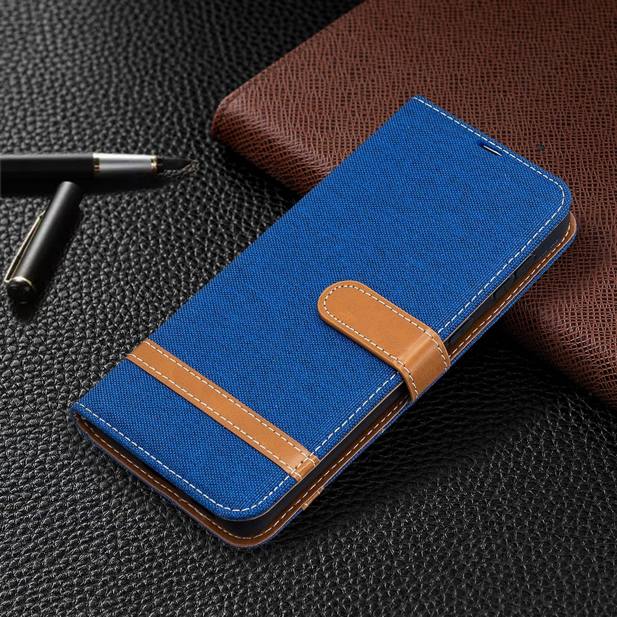 Blaue Handyhülle geschlossen mit braunem Riemen, auf strukturierter schwarzer Oberfläche, mit Stift.