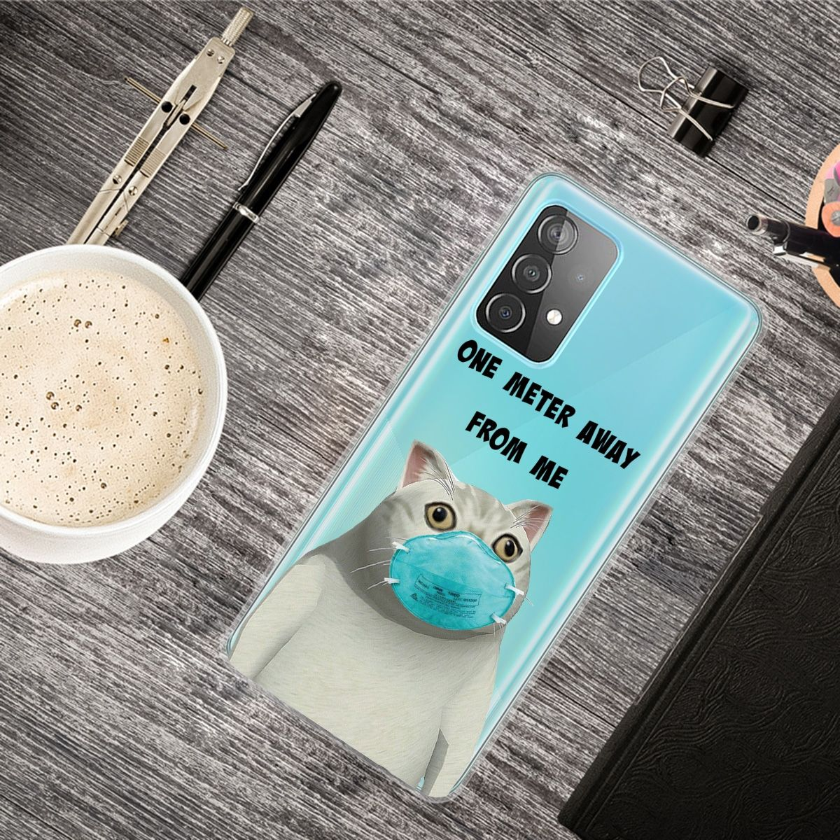 Eine Handyhülle mit einer Katze mit Maske. Es gibt Stifte und eine Tasse Kaffee.