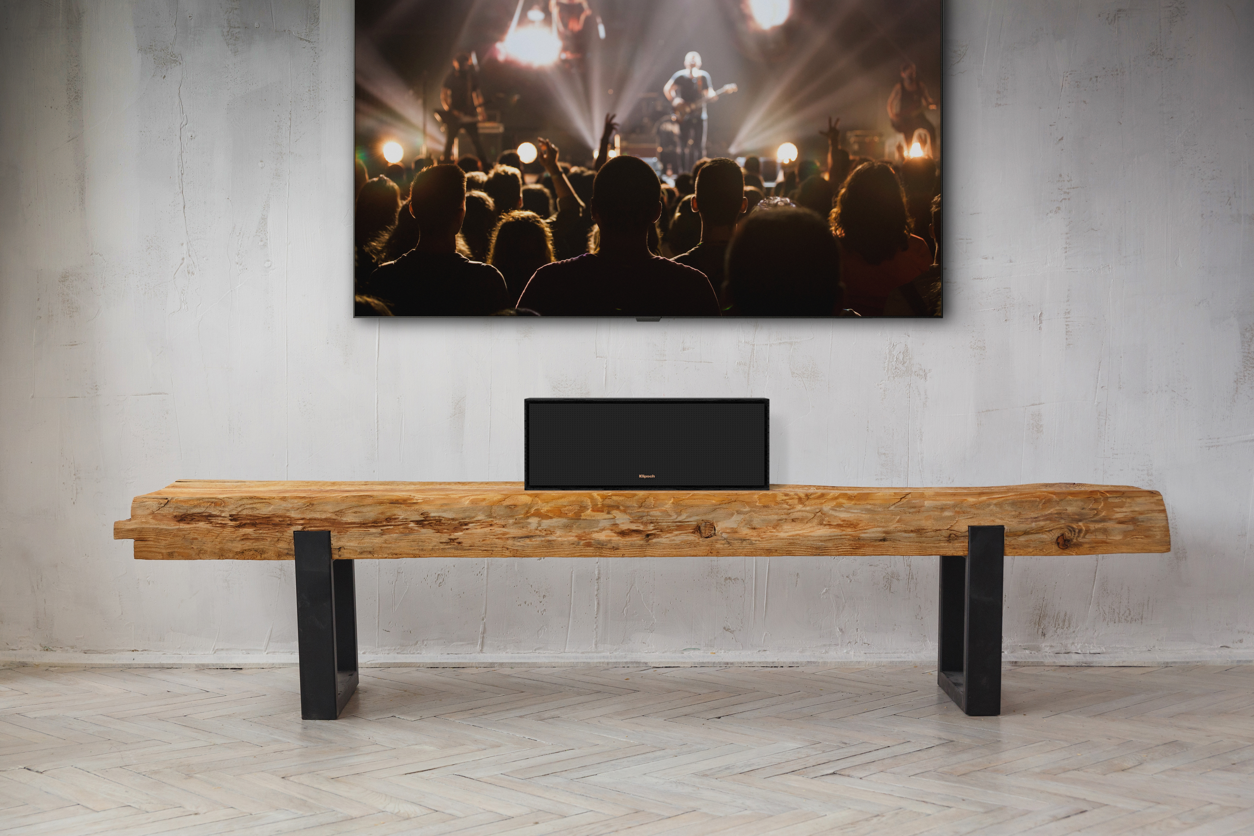 Ein schwarzer Lautsprecher steht auf einer Holzbank. Ein großer Fernseher zeigt eine Band vor Publikum.