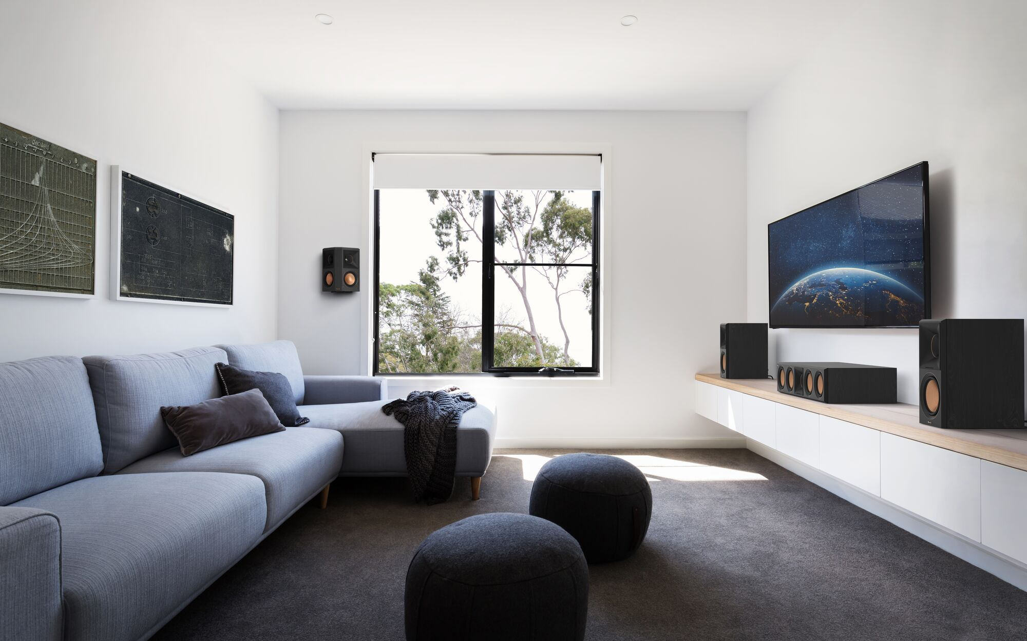 Een grijze bank in een moderne woonkamer met een grote tv, raam en luidsprekers.