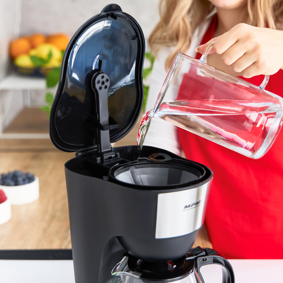 Kobieta wlewa wodę do czarnego ekspresu do kawy. Tło kuchni z owocami.