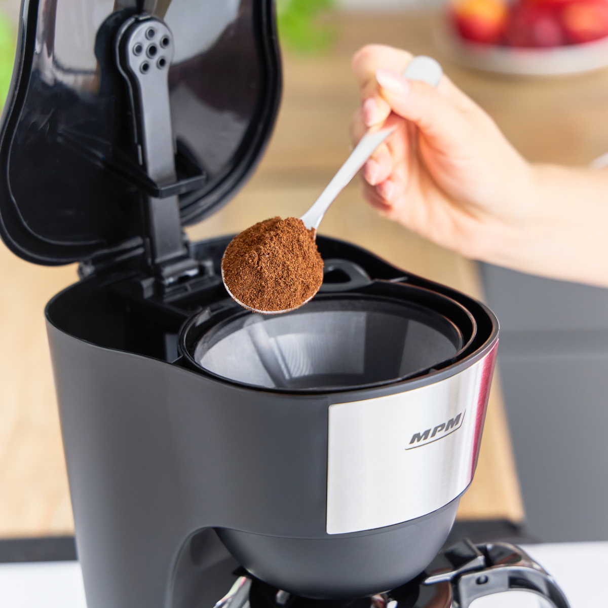 Ręka dodaje zmieloną kawę do czarnego ekspresu do kawy. Tło kuchni jest rozmyte.