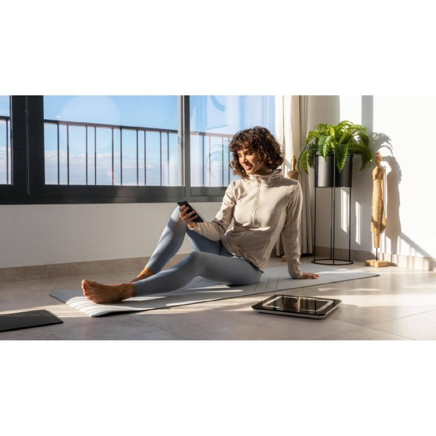 Mujer usando teléfono, sentada en esterilla yoga. Balanza y planta visibles.