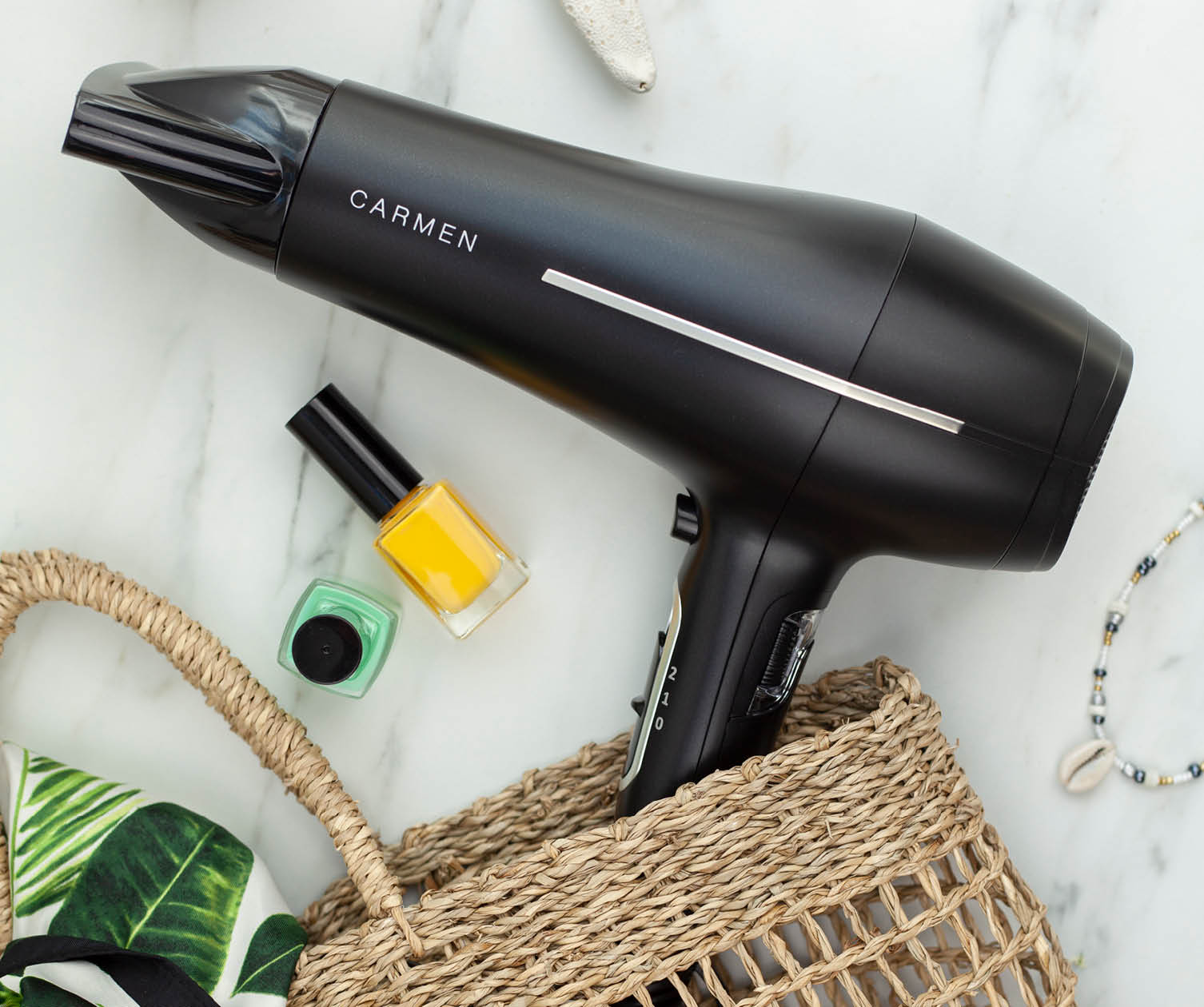 A black Carmen hair dryer in a straw basket, with nail polish and jewelry on a marble surface.