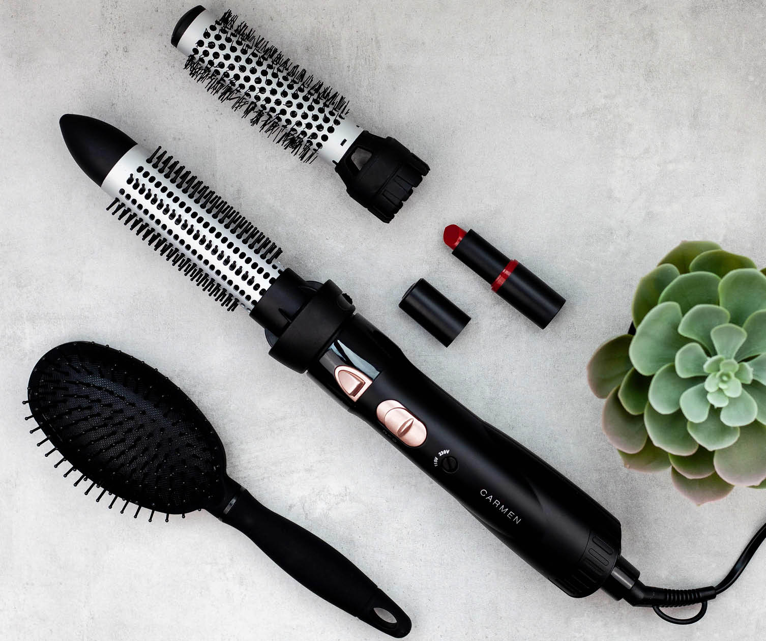 Strumenti per lo styling dei capelli, tra cui phon, spazzola e rossetto, su superficie grigia con succulenta.