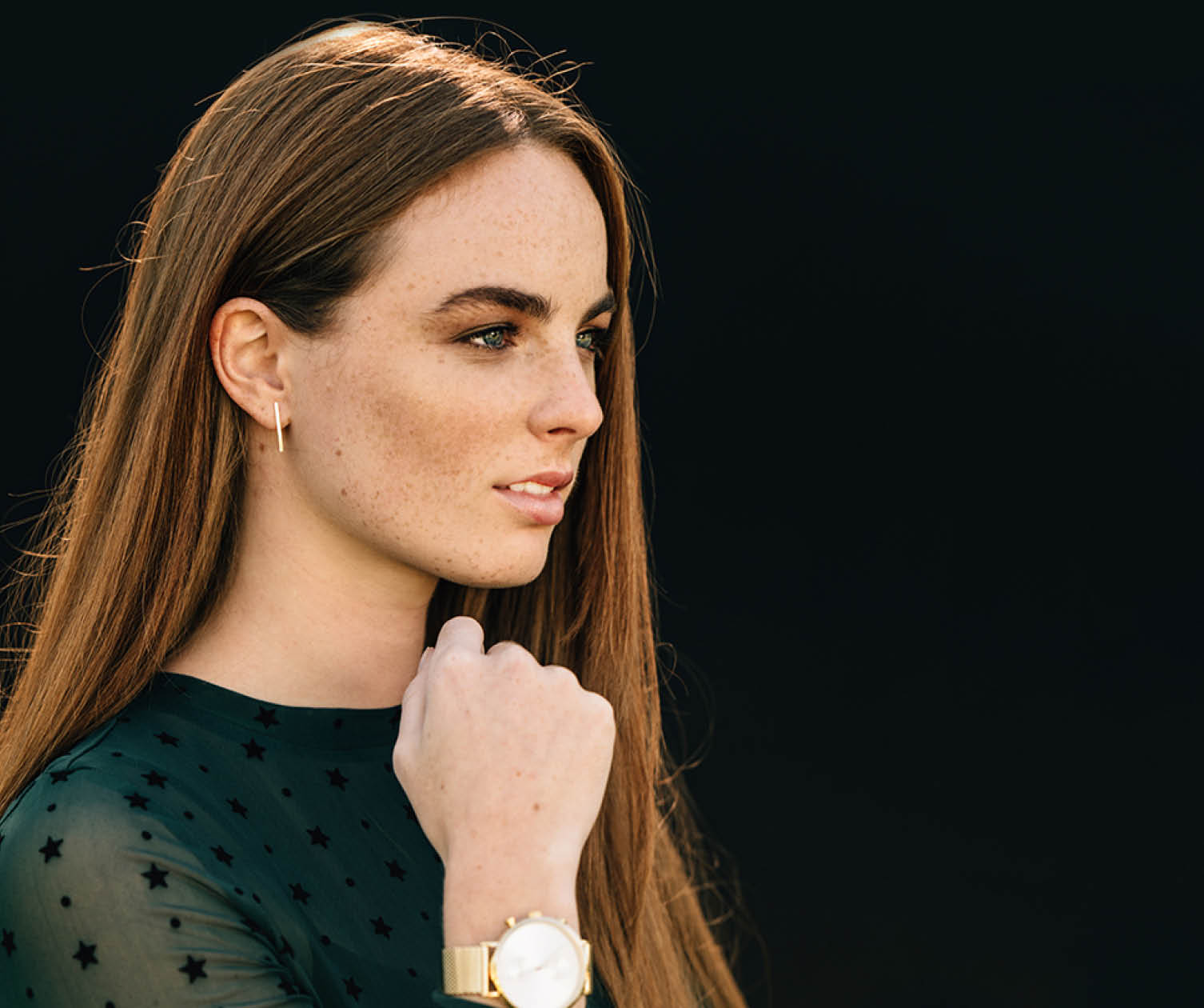 Donna con lentiggini con orologio e orecchino d'oro, guarda a sinistra su sfondo nero.