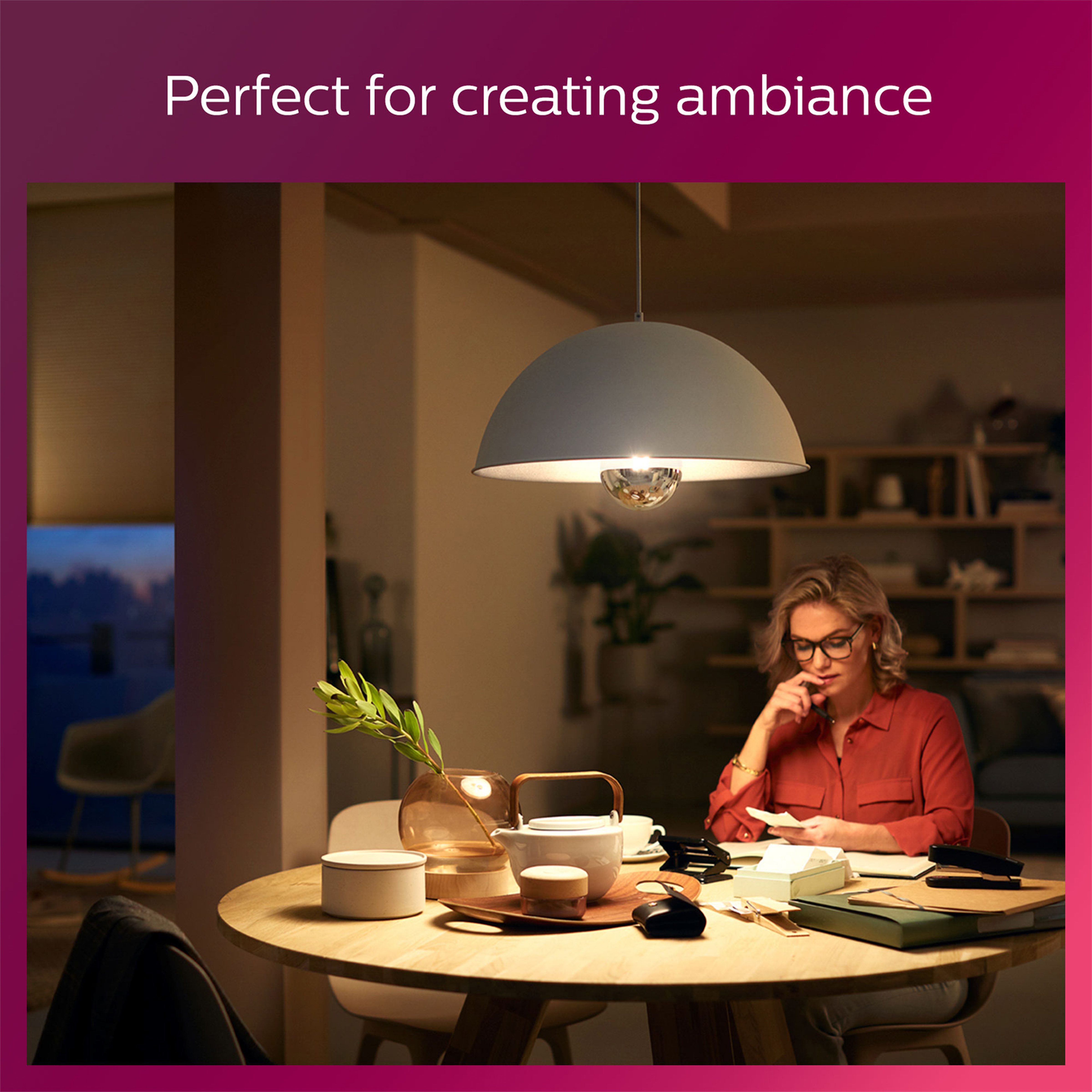 Frau sitzt an einem Tisch. Darüber hängt eine Lampe. Die Szene ist drinnen.