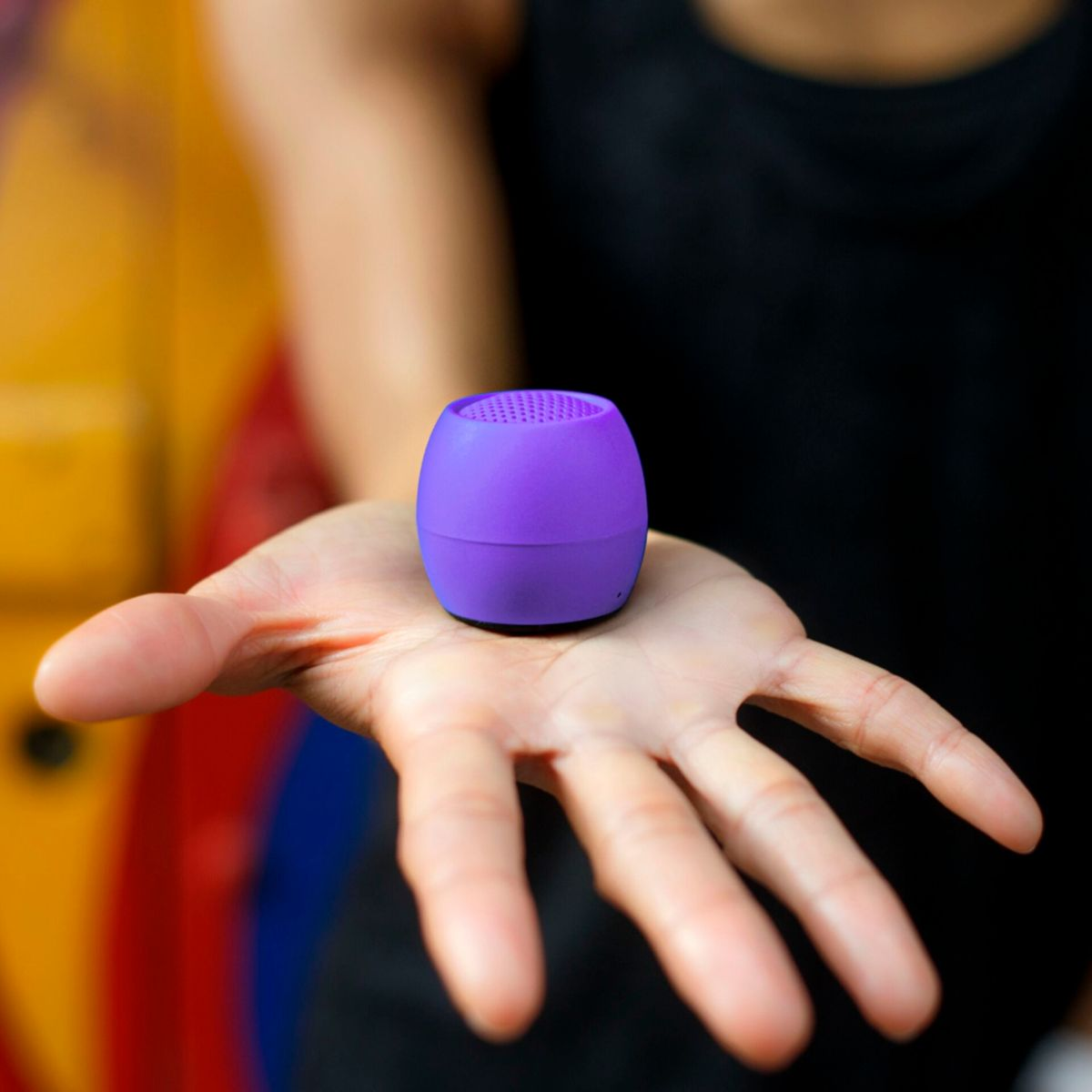 Fioletowy głośnik w kształcie jaja trzymany w otwartej dłoni osoby, na kolorowym tle.