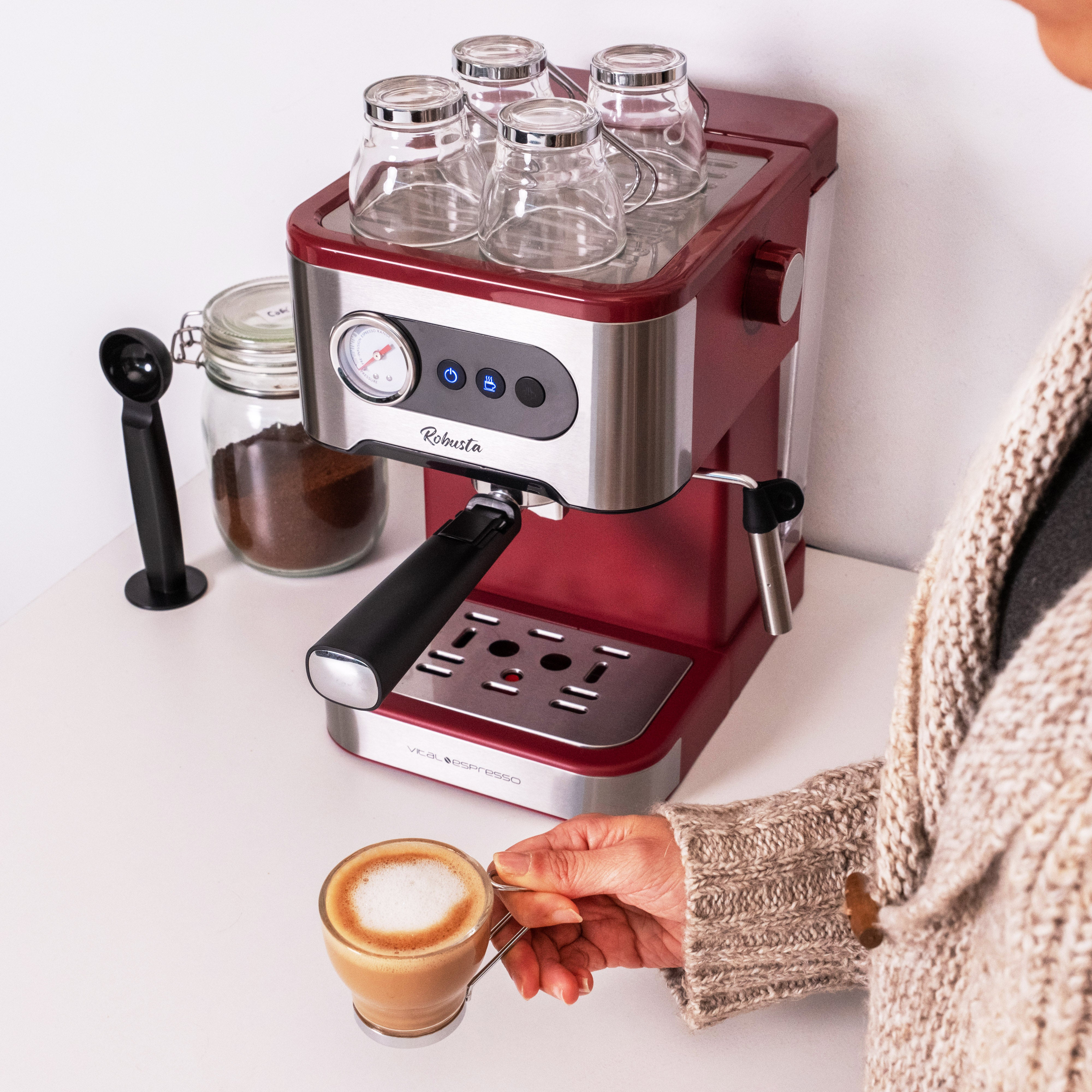 Mano de mujer sosteniendo un vaso de capuchino. Una máquina de espresso y un tarro de café están cerca.