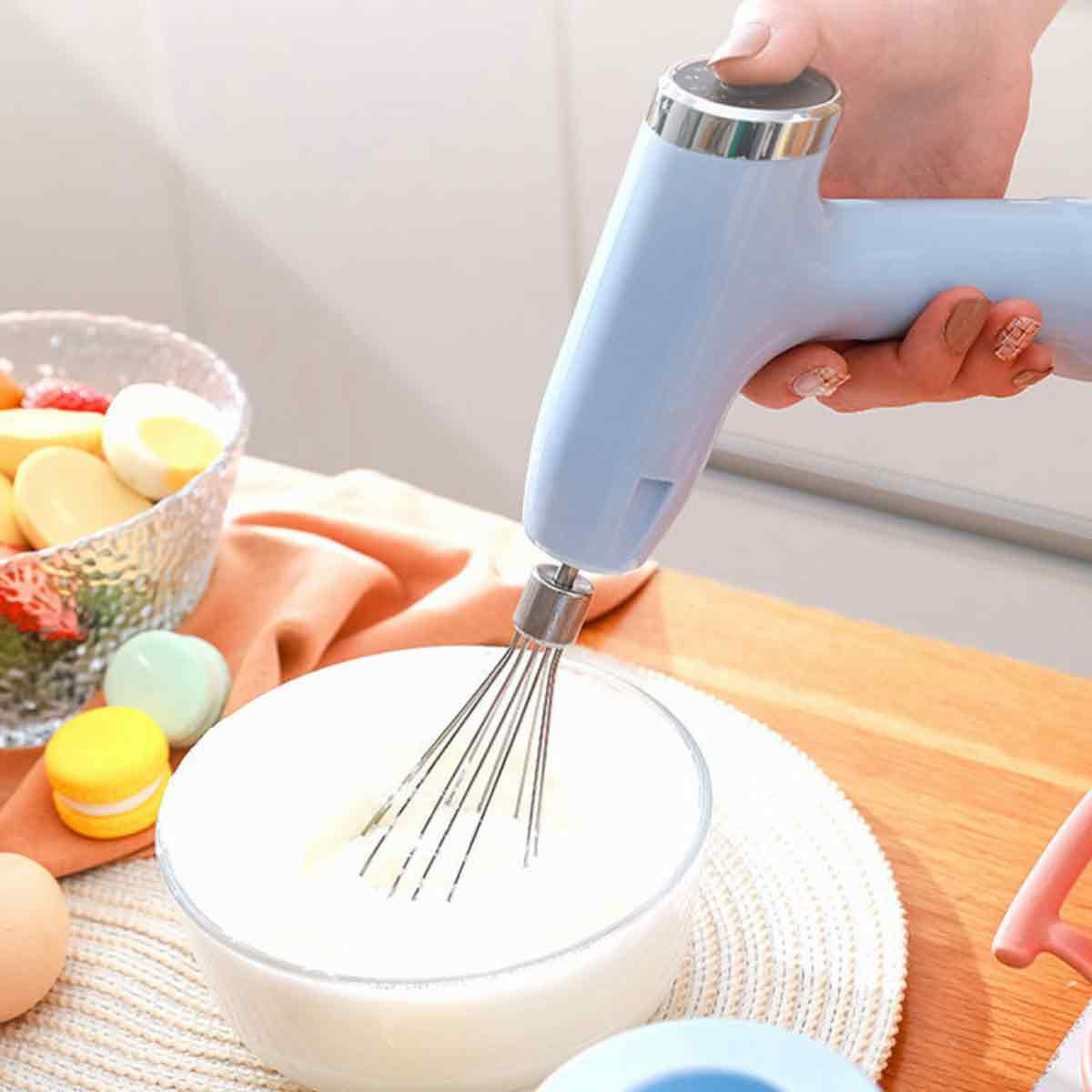 Hand hält einen blauen elektrischen Mixer. Eine Schüssel mit Teig und Essen auf dem Tisch.