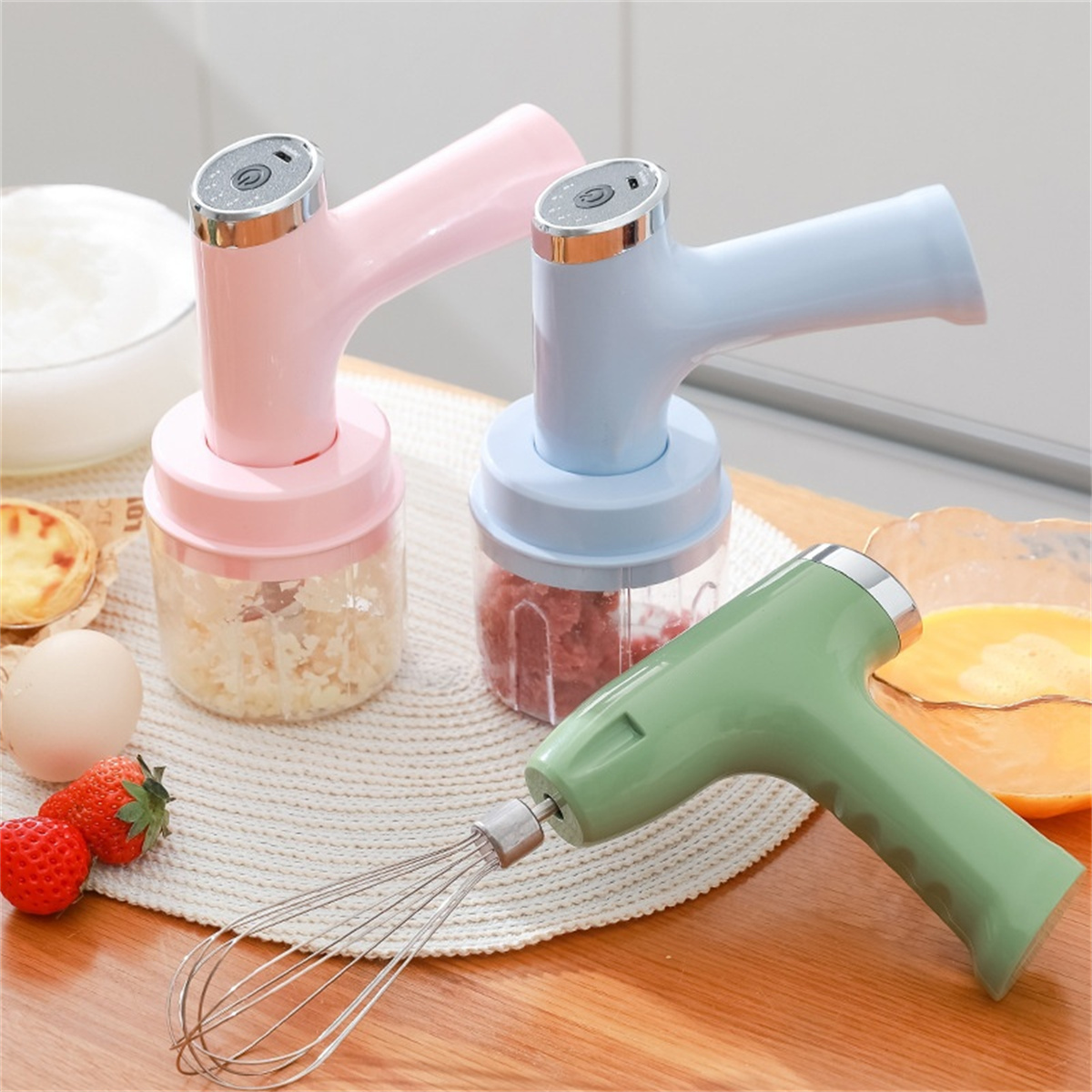 Drei elektrische Mixer, rosa, blau und grün, mit Zutaten und Lebensmitteln im Hintergrund.