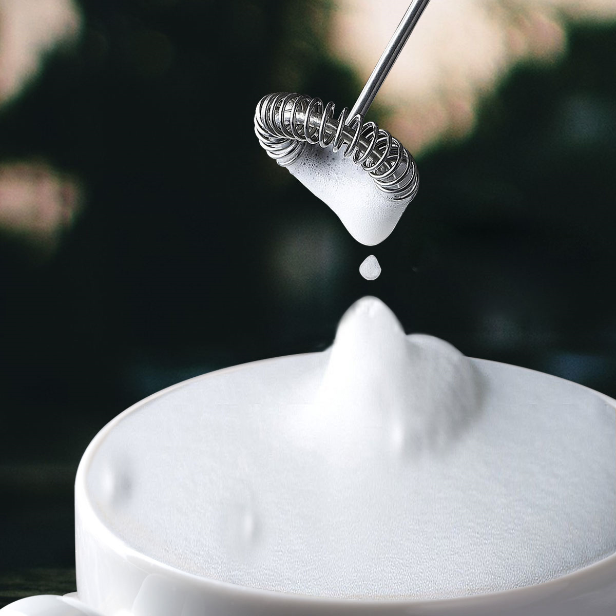 Eine weiße Tasse mit schaumiger Milch gefüllt. Ein Milchaufschäumer wird darüber gehalten, tropft Milch auf den Schaum.