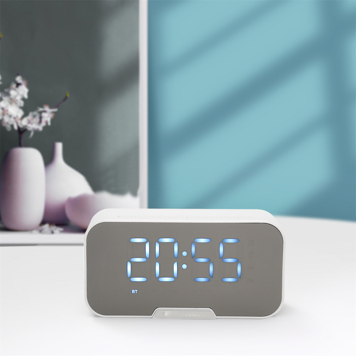 Weißer Digitalwecker mit der Anzeige 20:55 auf einem Tisch. Dahinter weiße Vasen und eine türkisfarbene Wand.