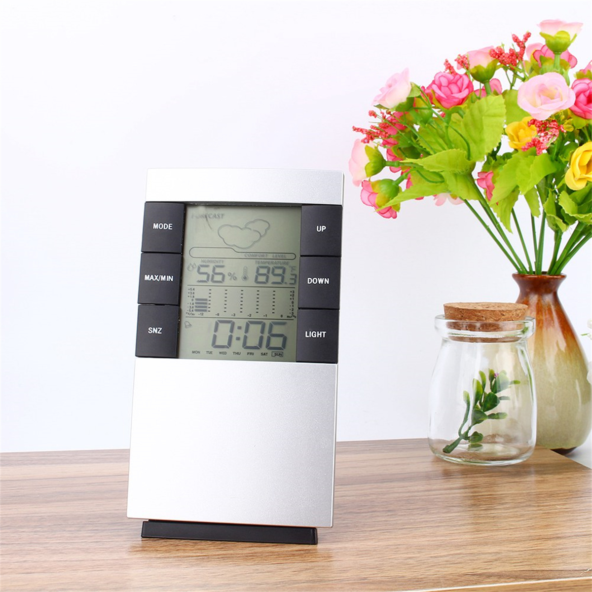 Silberne Wetterstation mit Anzeige von Luftfeuchtigkeit, Temperatur und Uhrzeit. Blumen im Hintergrund.