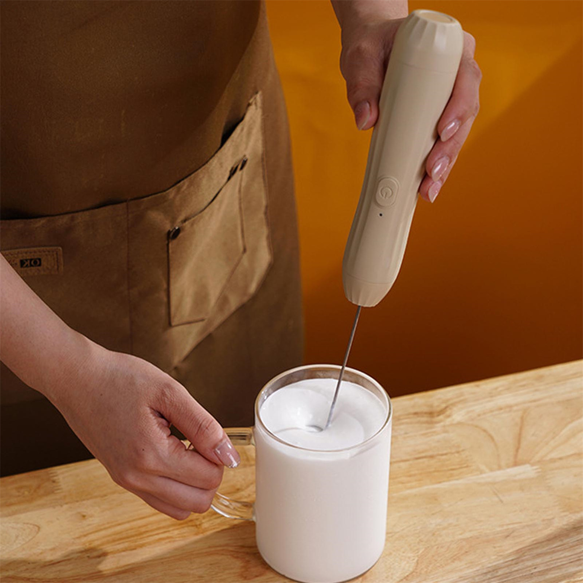 Eine Hand hält einen Milchaufschäumer über einem Glas Milch auf einer Holzoberfläche.