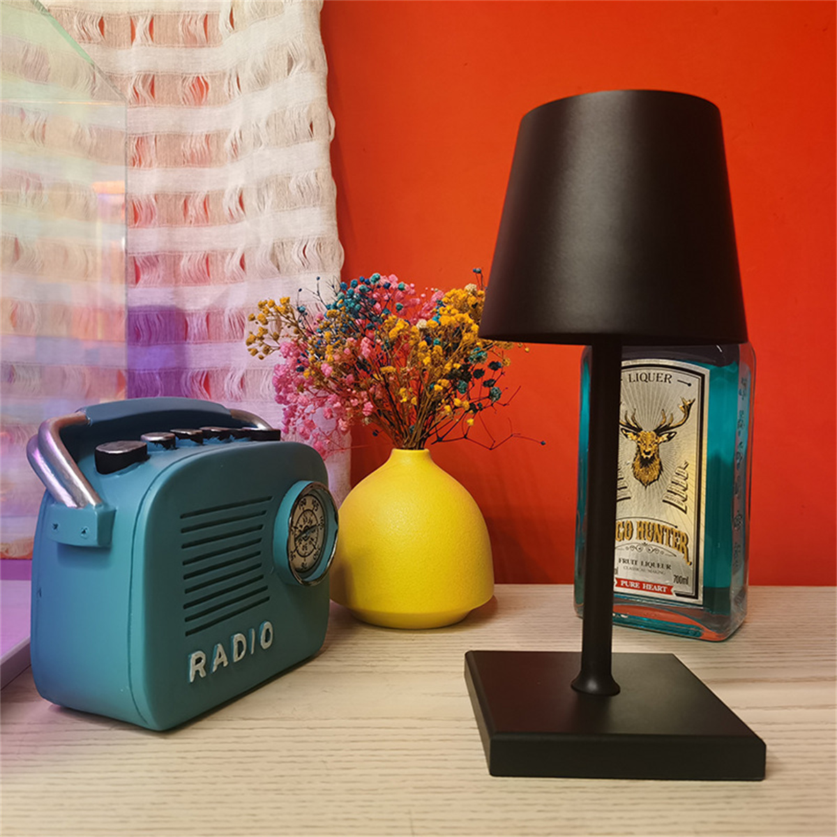 Eine schwarze Lampe mit schwarzem Schirm, neben einem blauen Radio und einer gelben Vase mit Blumen.