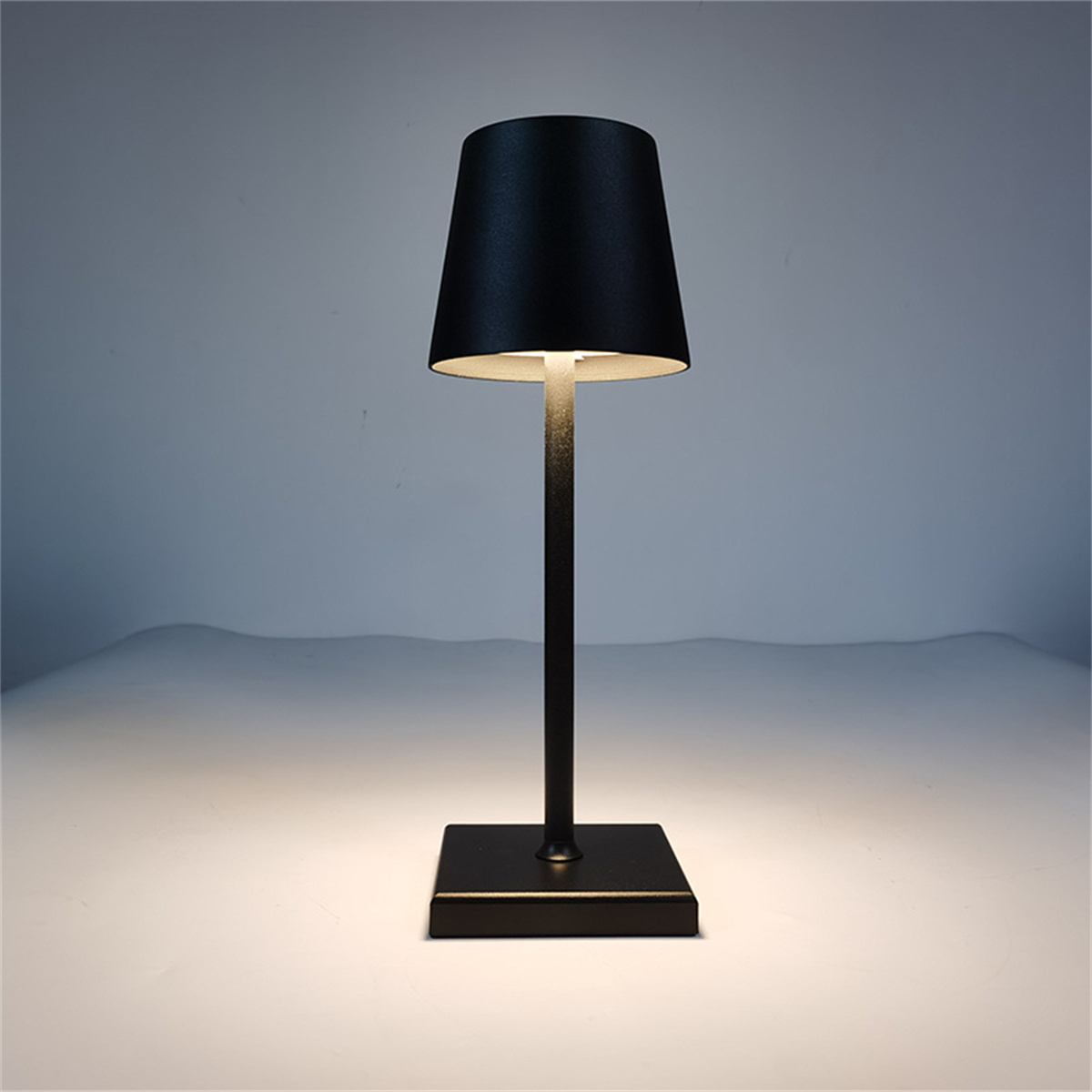 Schwarze Tischlampe, quadratischer Sockel und dünner Pfahl, mit schwarzem Lampenschirm. Der Hintergrund ist weiß.