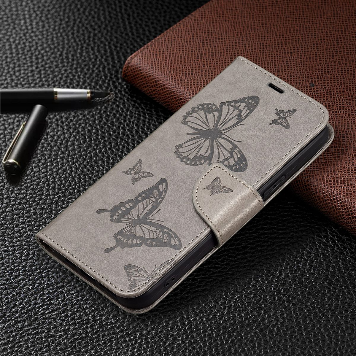 Graue Handyhülle mit Schmetterlingsdesign, teilweise geöffnet auf dunkler Oberfläche mit Stift und Lederdetails.
