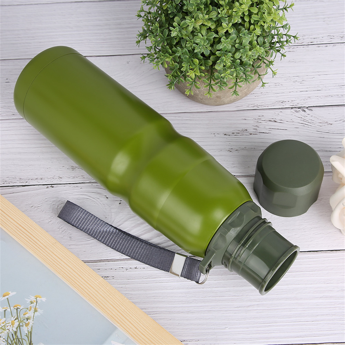 Grüne Wasserflasche mit Riemen und Deckel. Pflanze und Holz-Bilderrahmen im Hintergrund.