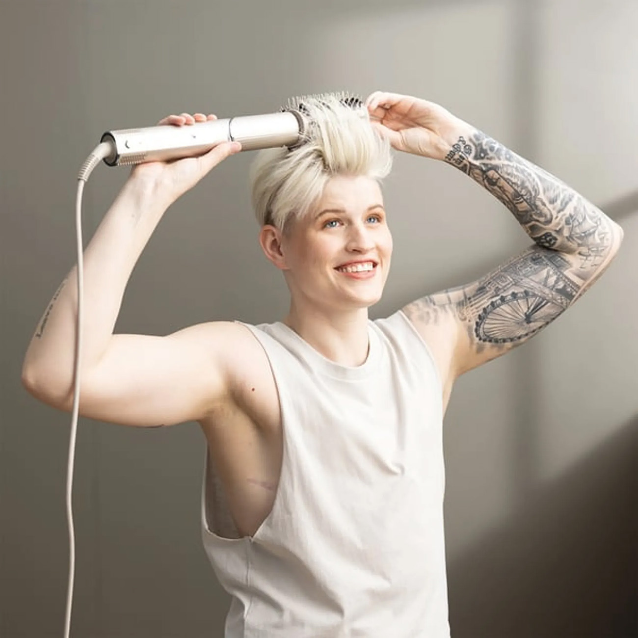 Kobieta z krótkimi, blond włosami, stylizująca je szczotką do włosów. Ma tatuaż na ramieniu.