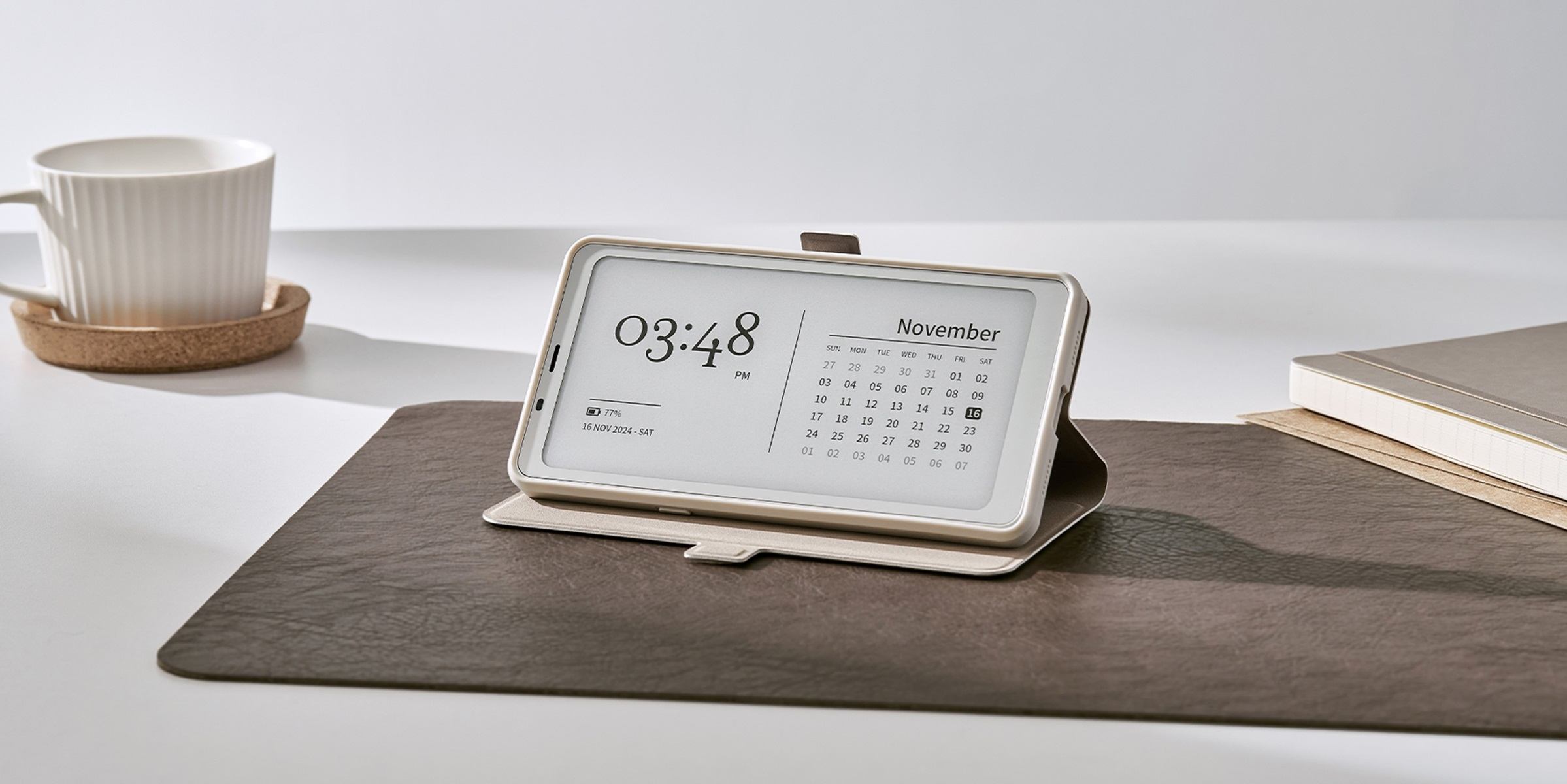E-reader su scrivania mostra ora e calendario. Tazza e quaderno visibili sullo sfondo.