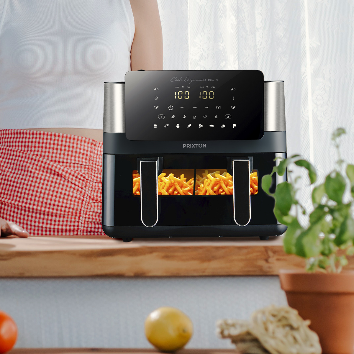 Frytkownica powietrzna z frytkami w środku. Kobieta stoi za nią. Na drewnianej powierzchni z rośliną.