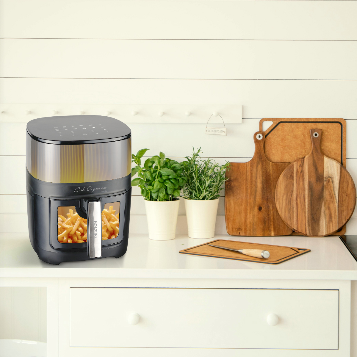 Frytownica powietrzna z frytkami w środku, obok roślin, desek do krojenia i otoczenia kuchennego.