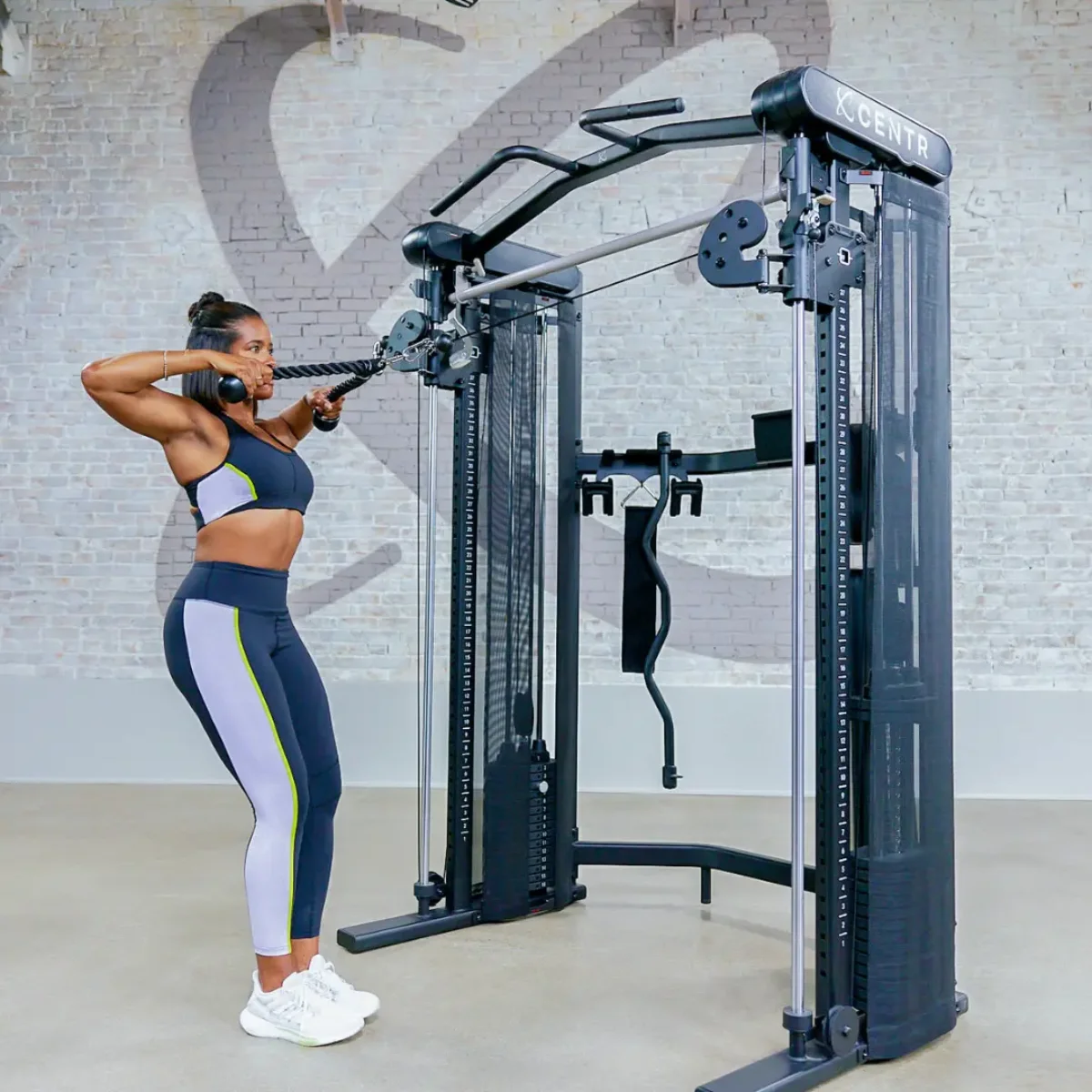 Frau, die eine Kabelmaschine benutzt. Sie trainiert, mit Fitnessgeräten und einer Backsteinmauer im Hintergrund.
