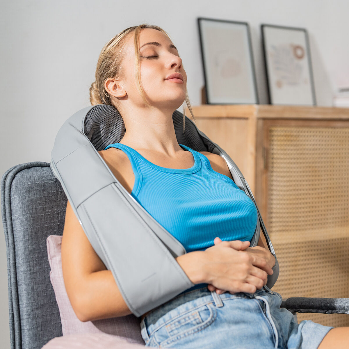 Mujer relajándose con un masajeador de cuello gris, ojos cerrados, top azul y pantalones cortos de mezclilla.